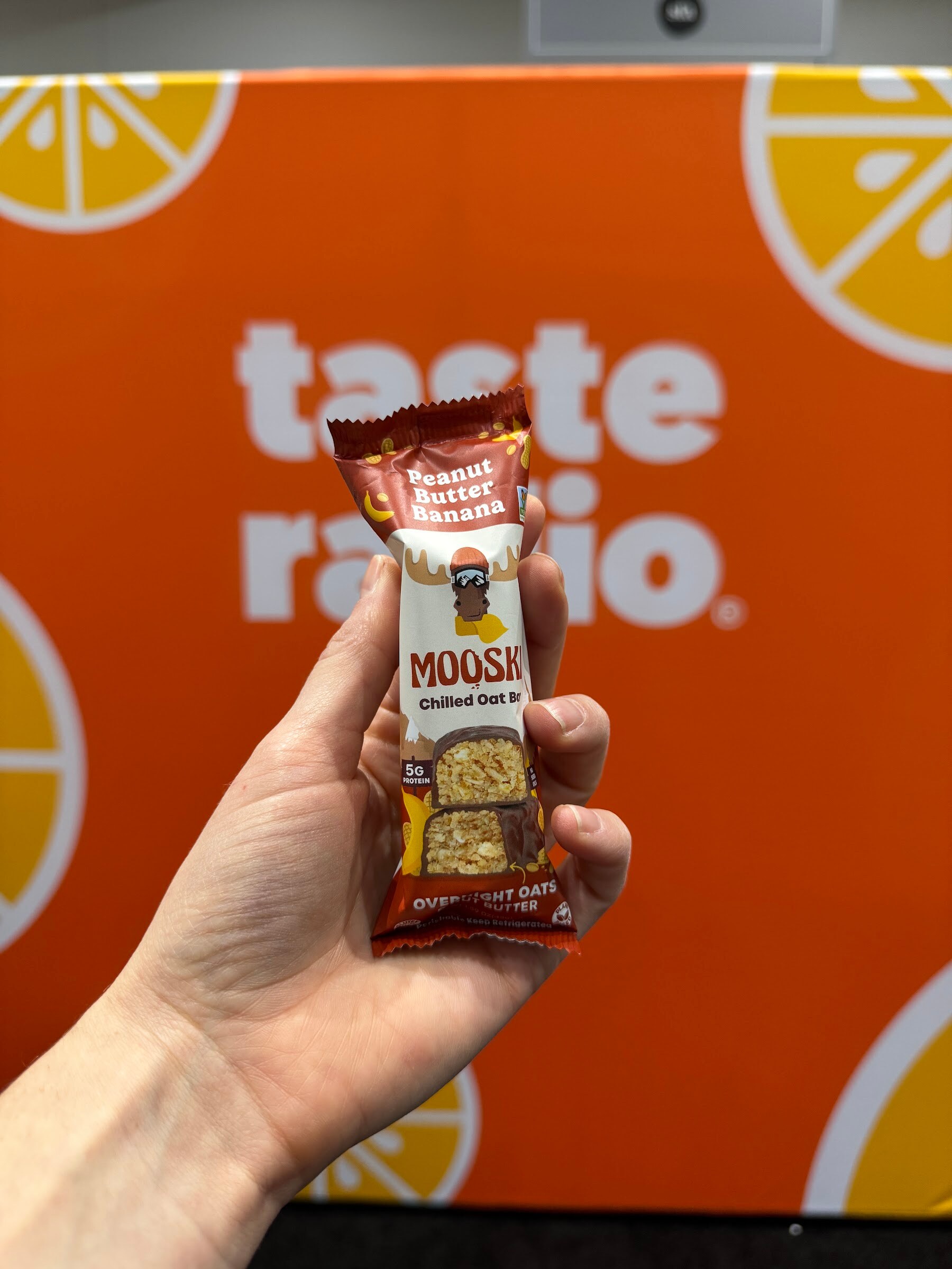 Hand holds a Mooski peanut butter banana chilled oat bar in front of the Taste Radio backdrop.