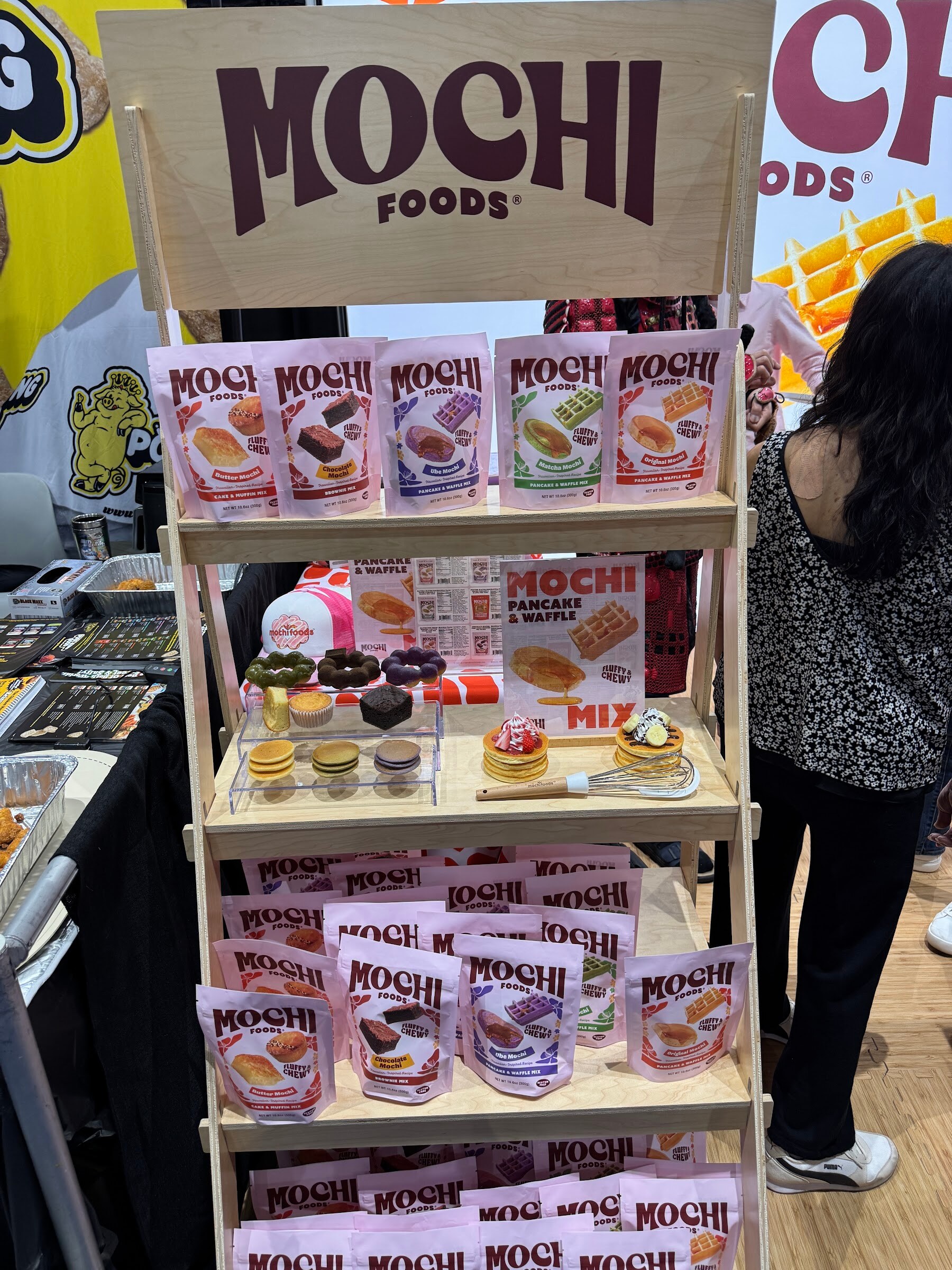 Wooden shelf display topped with a Mochi Foods sign holding pink and purple mochi mix bags.