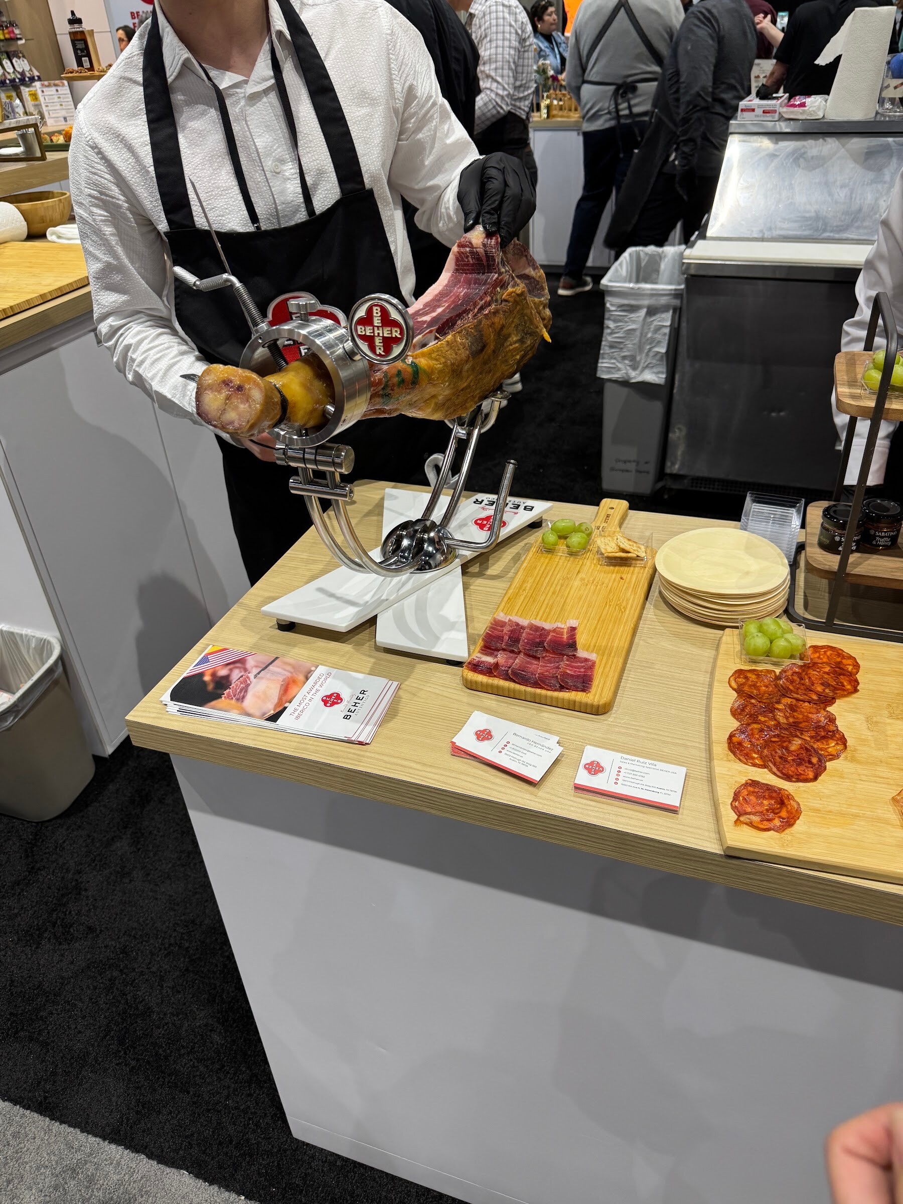 Server in a black apron carves a leg of cured ham on a stand beside a board of sliced samples.