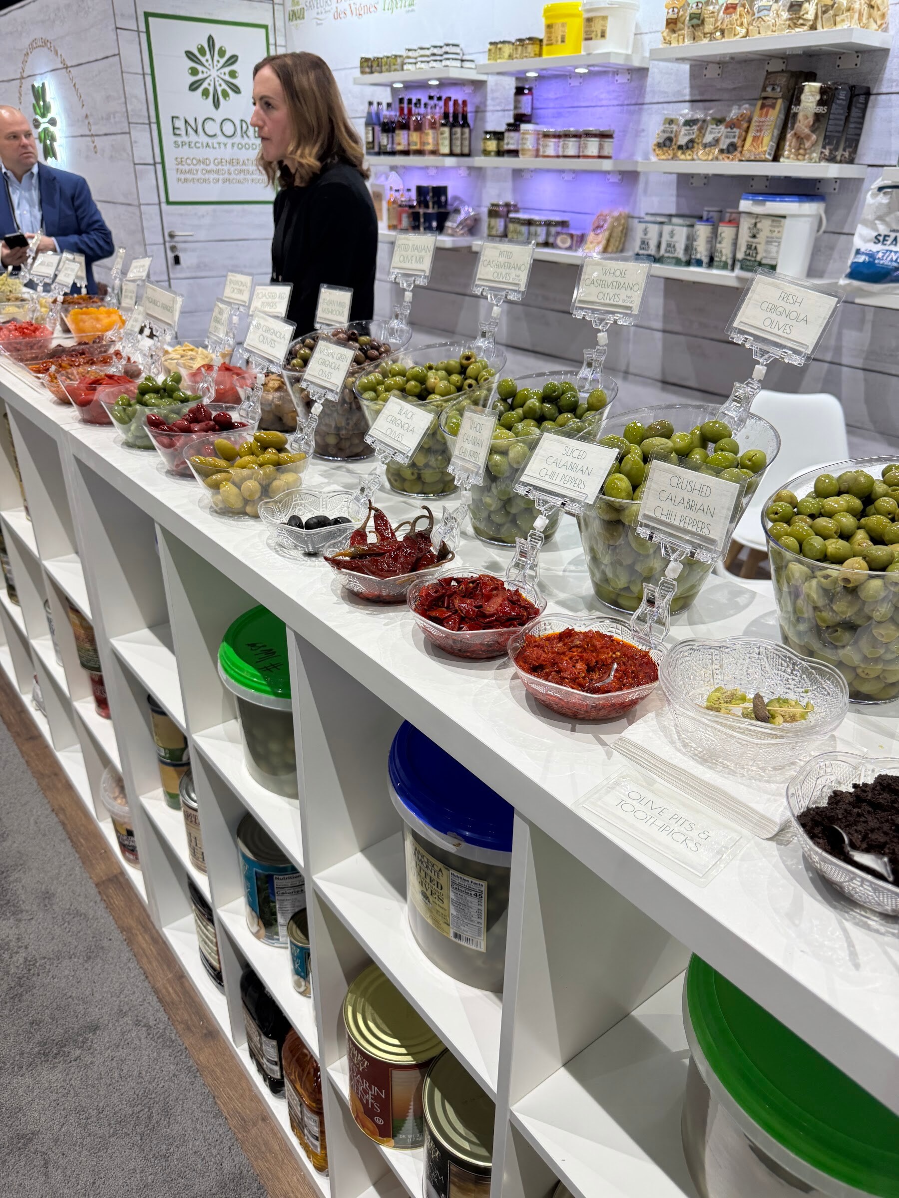 Long white counter lined with bowls of olives, peppers and antipasti in front of shelves of jars.