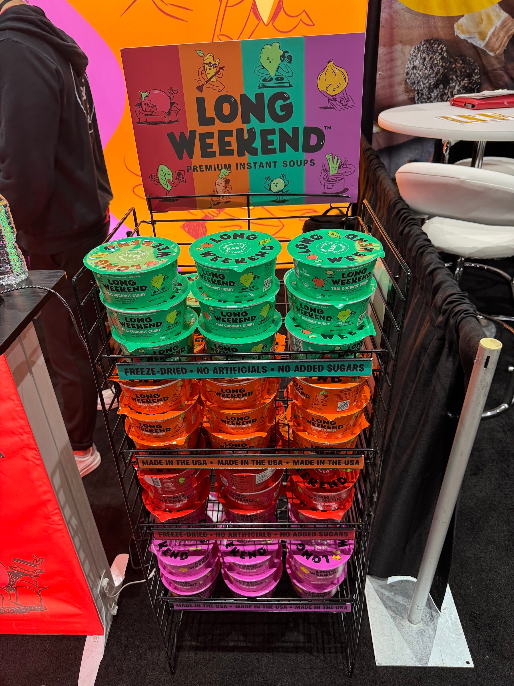 Wire rack stacked with green, orange and pink cups of Long Weekend instant soup under a colorful sign.