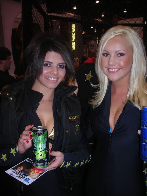 Two smiling women in black Rockstar outfits with gold stars pose together; one holds a green Rockstar 21 can, and the other holds a blue can. A chain-link cage is behind them.
