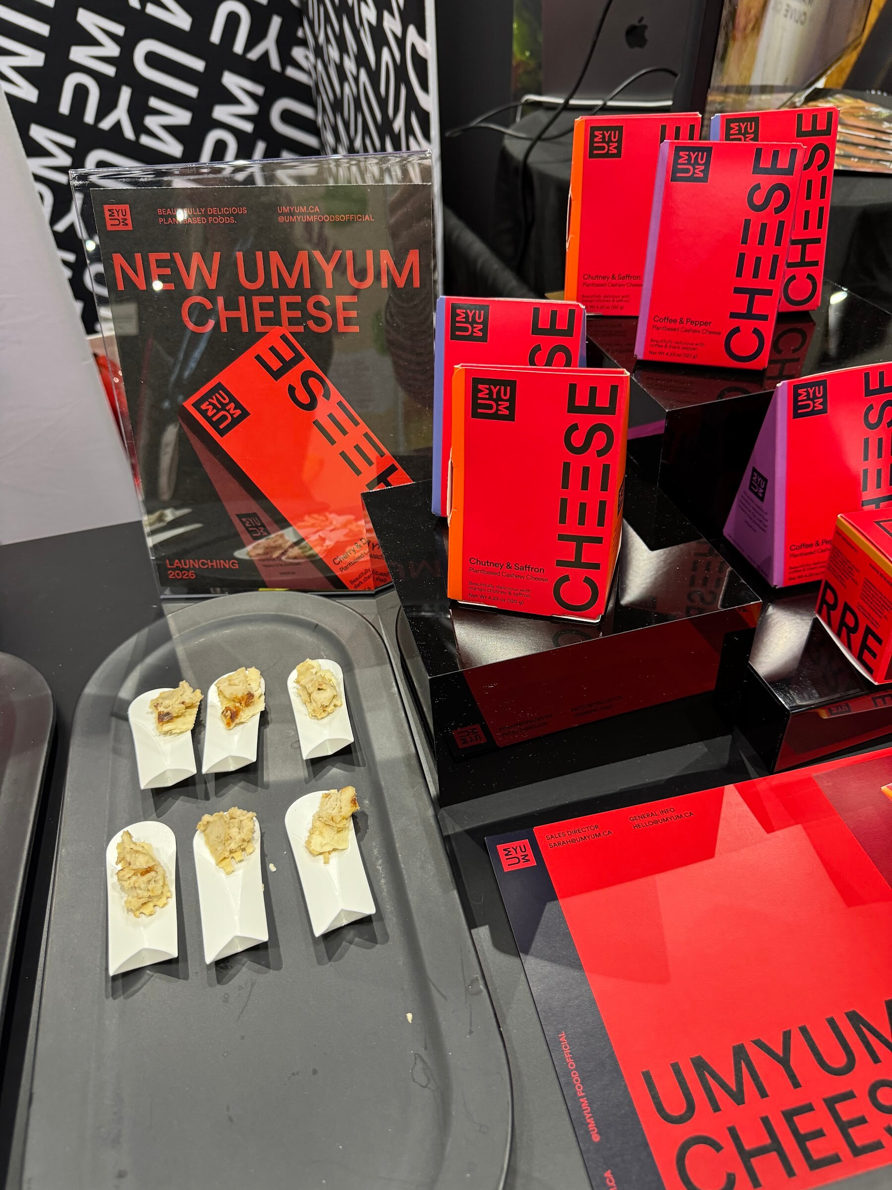 Red UMYUM cheese boxes on black risers behind a tray of cheese samples on crackers.