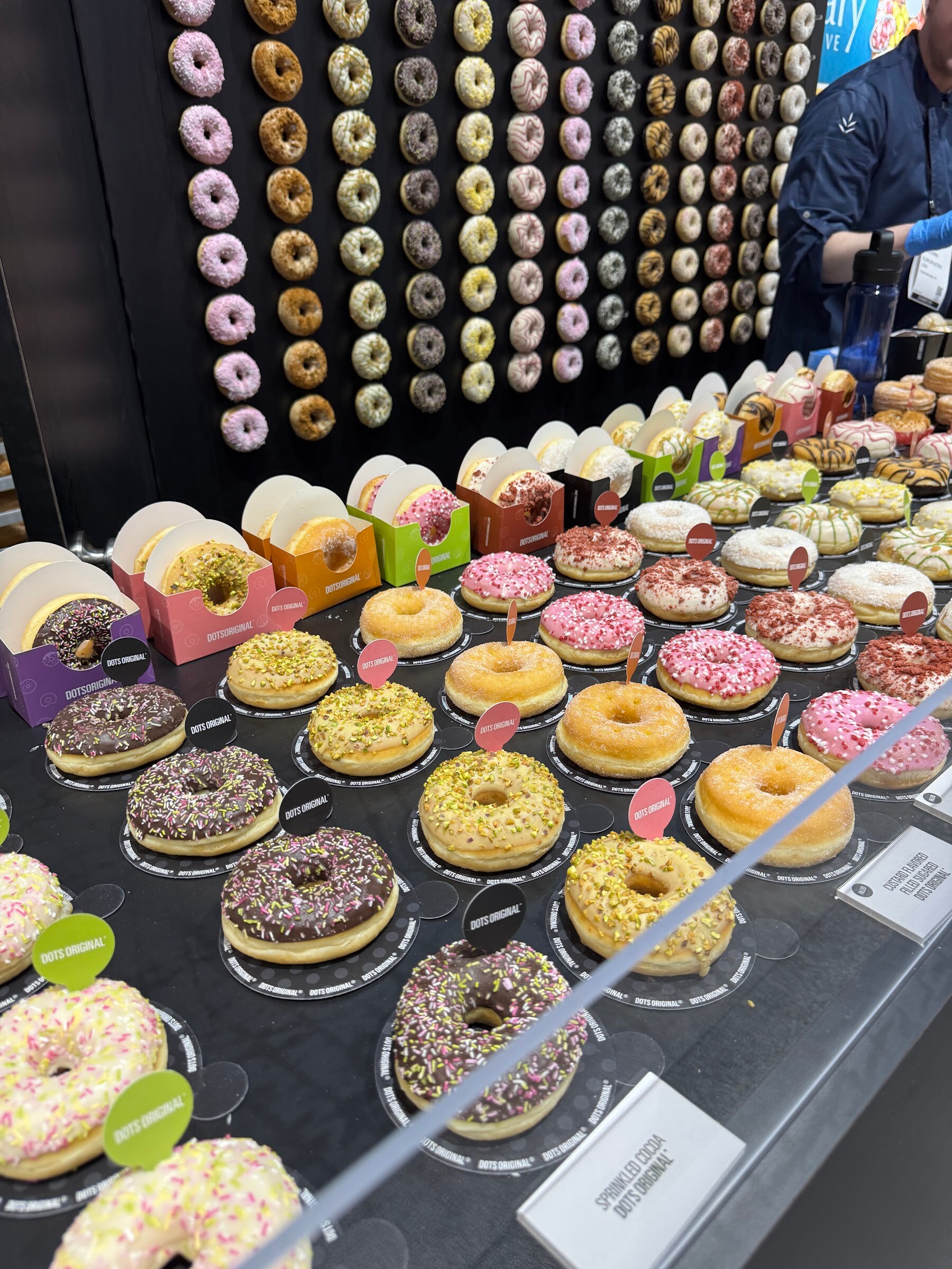 Rows of colorful glazed and sprinkled donuts on a black counter in front of a donut-covered wall.