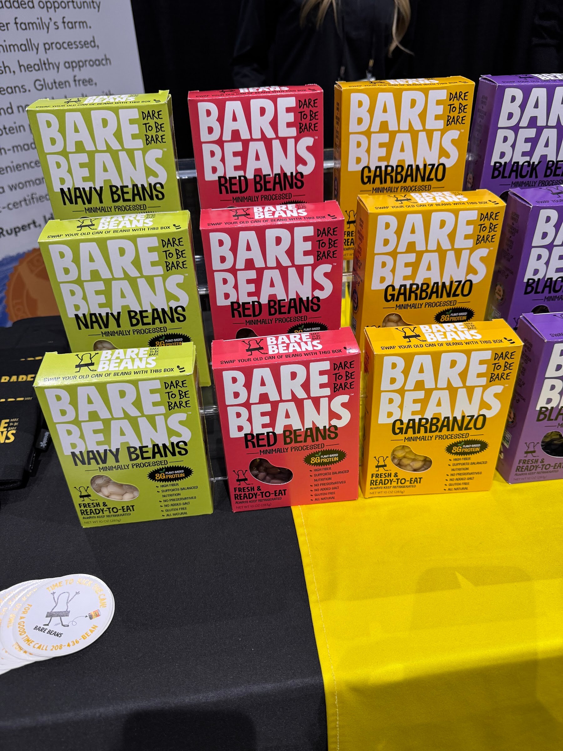 Stacked boxes of Bare Beans in green, red, yellow and purple on a black and yellow table.