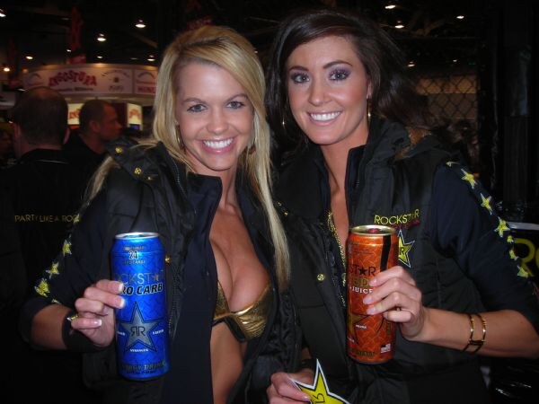 Two smiling women in black Rockstar jackets hold cans: one a blue Rockstar Zero Carb, the other an orange Rockstar Juiced, with an expo hall behind them.