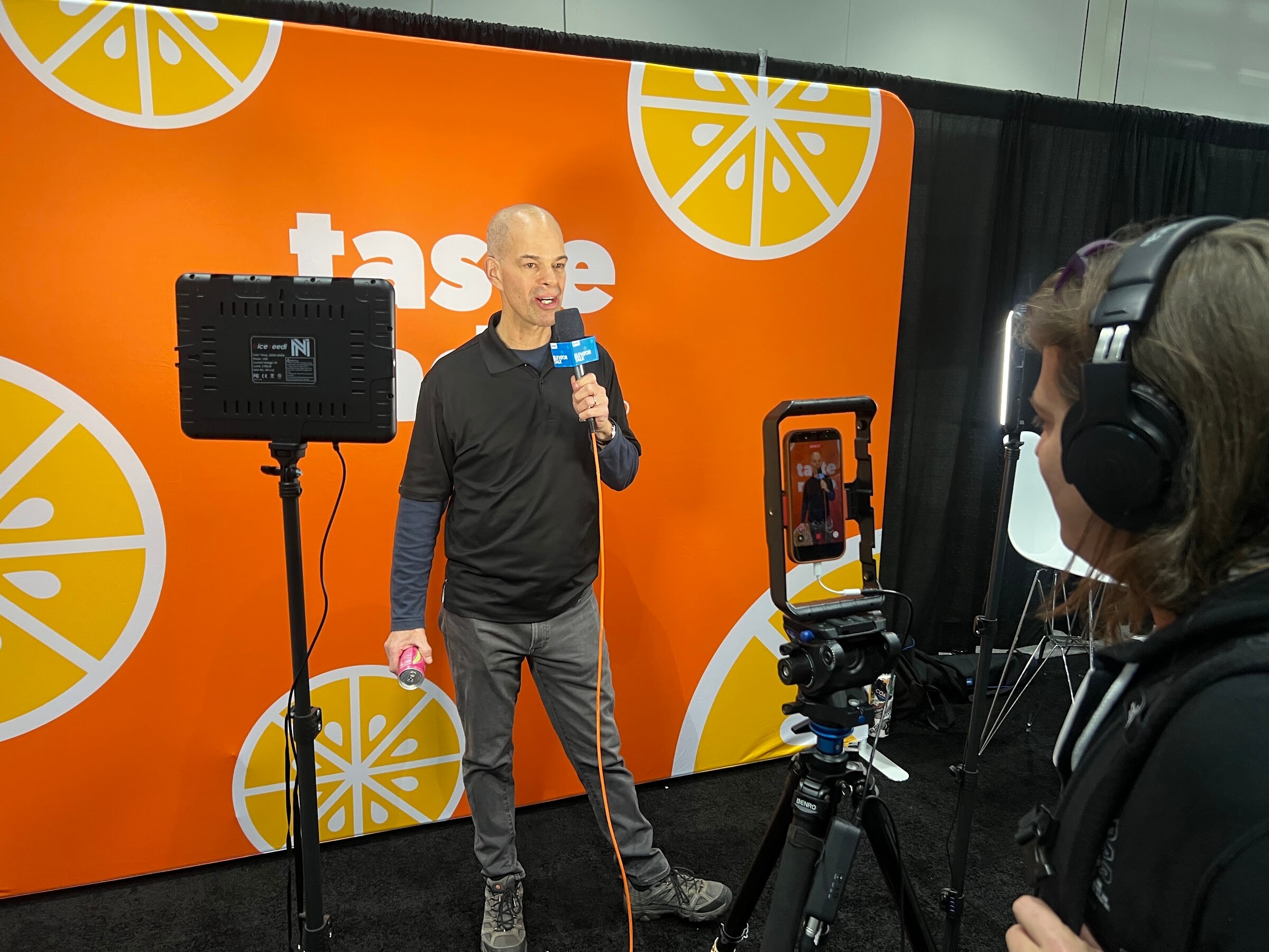 Man holding a microphone stands before an orange Taste Radio backdrop while a camera operator in headphones films with a phone rig and lights.