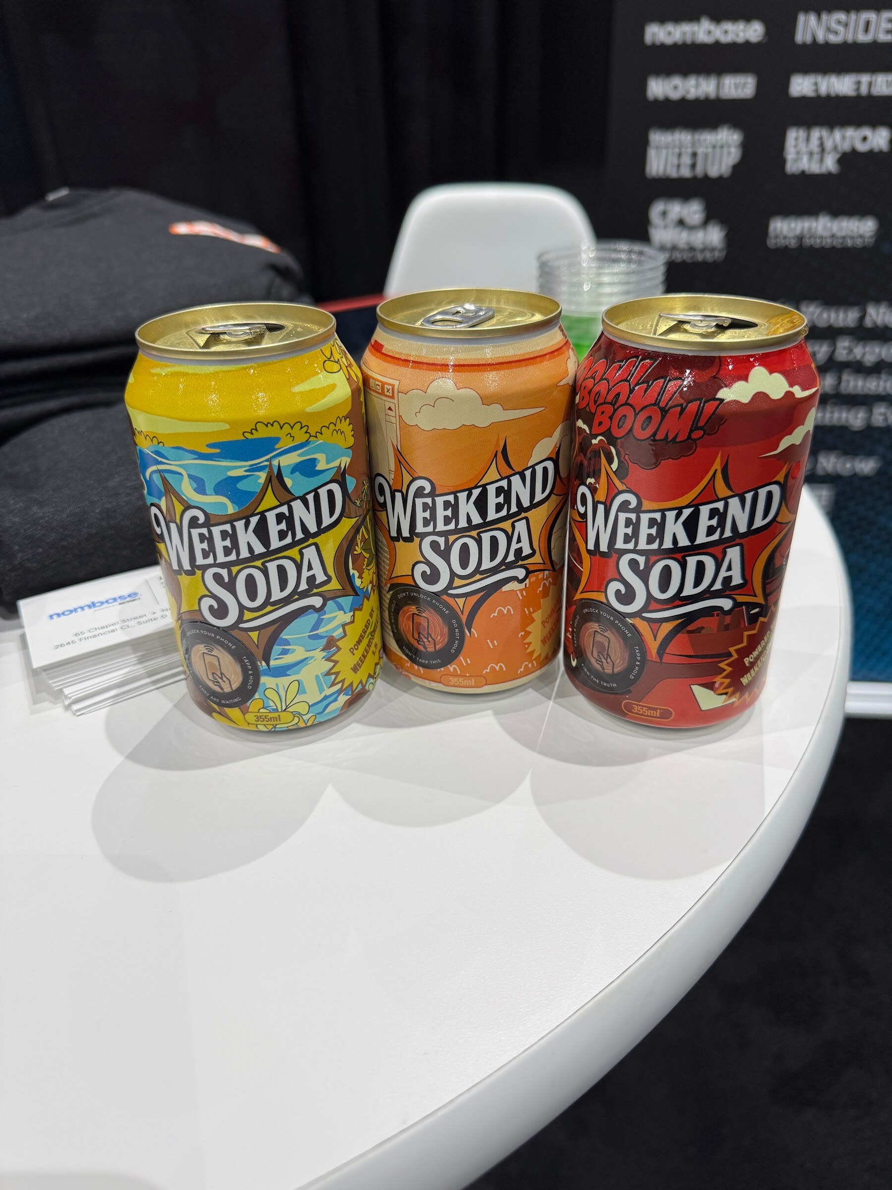 Three cans of Weekend Soda on a white table in front of a backdrop with Nosh, BevNET and Nombase logos.