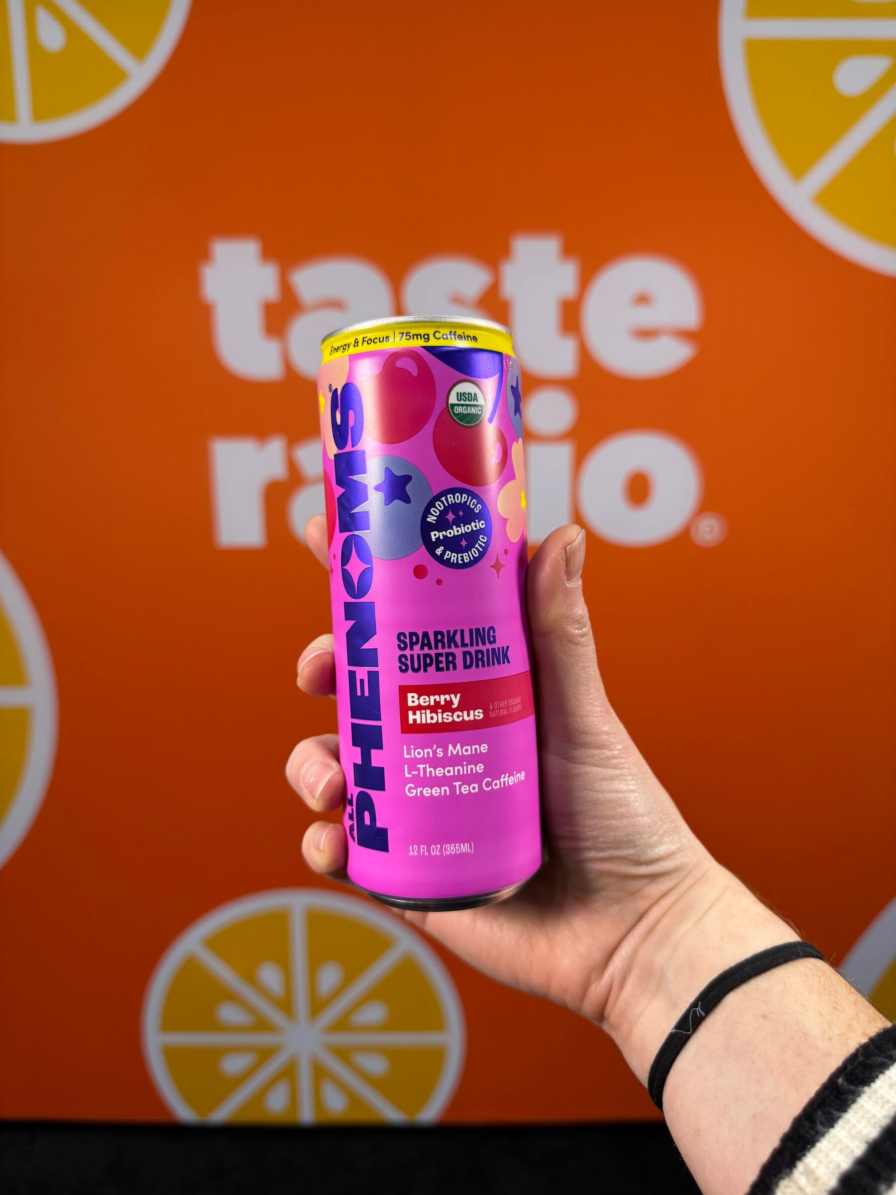 Hand holds a pink can of Phenoms Berry Hibiscus sparkling super drink in front of the Taste Radio backdrop.