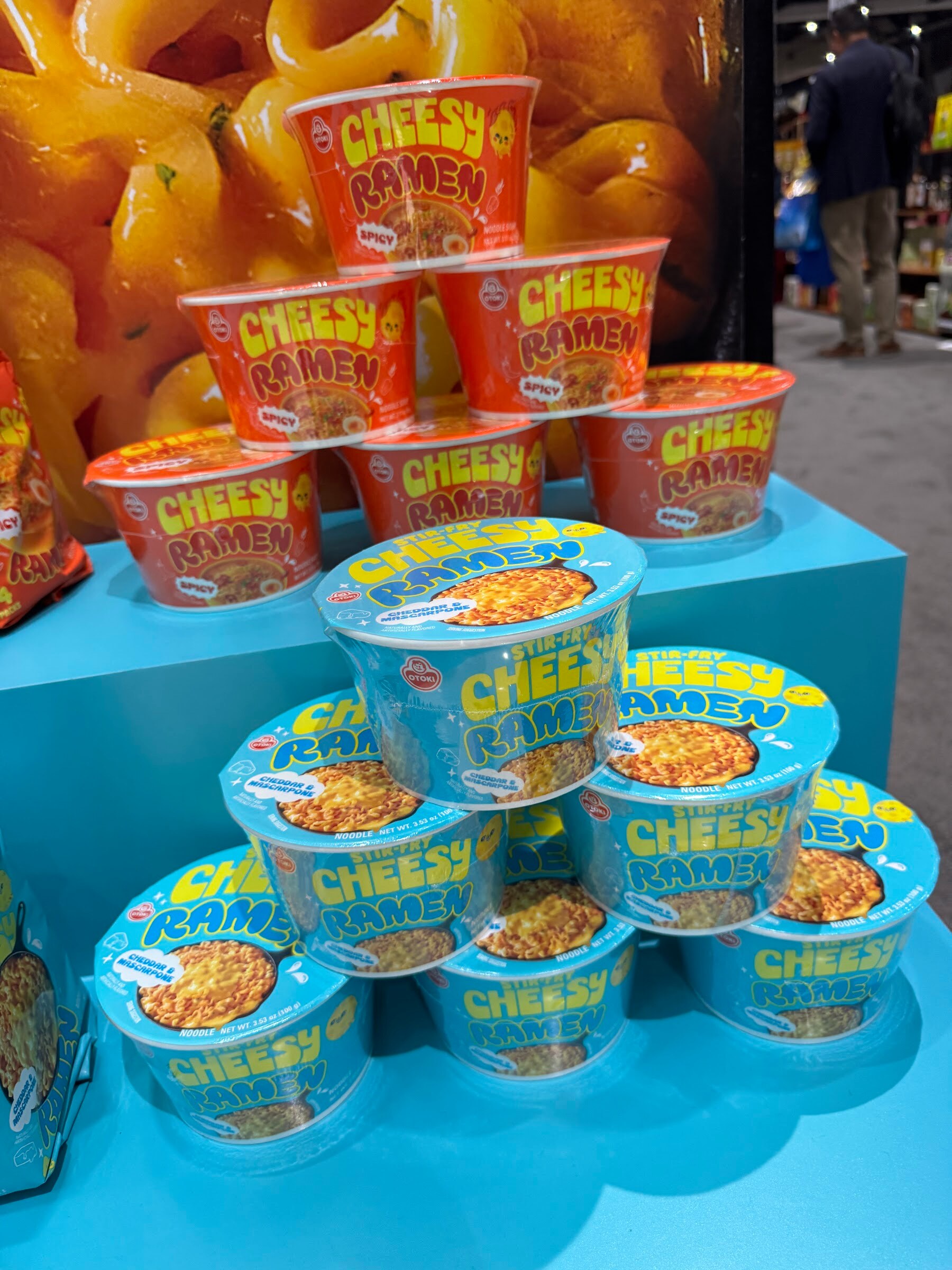 Stacked orange and blue Cheesy Ramen cups on a turquoise display.