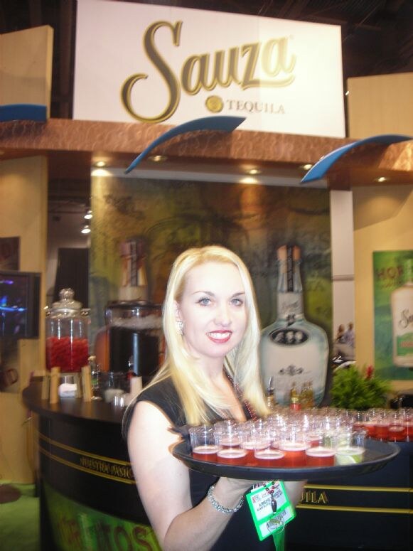 A smiling blonde woman holds a tray of small red shot cups in front of a booth with a large Sauza Tequila sign and a Sauza bottle graphic.