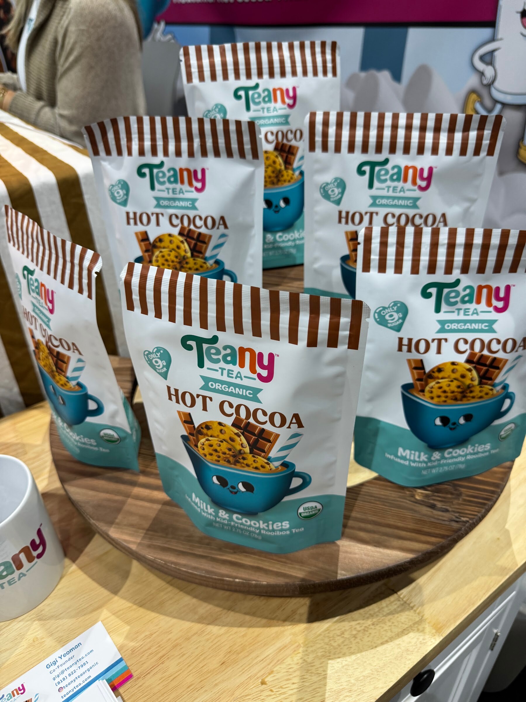 Pouches of Teany Tea organic hot cocoa with a cartoon mug on a wooden display.