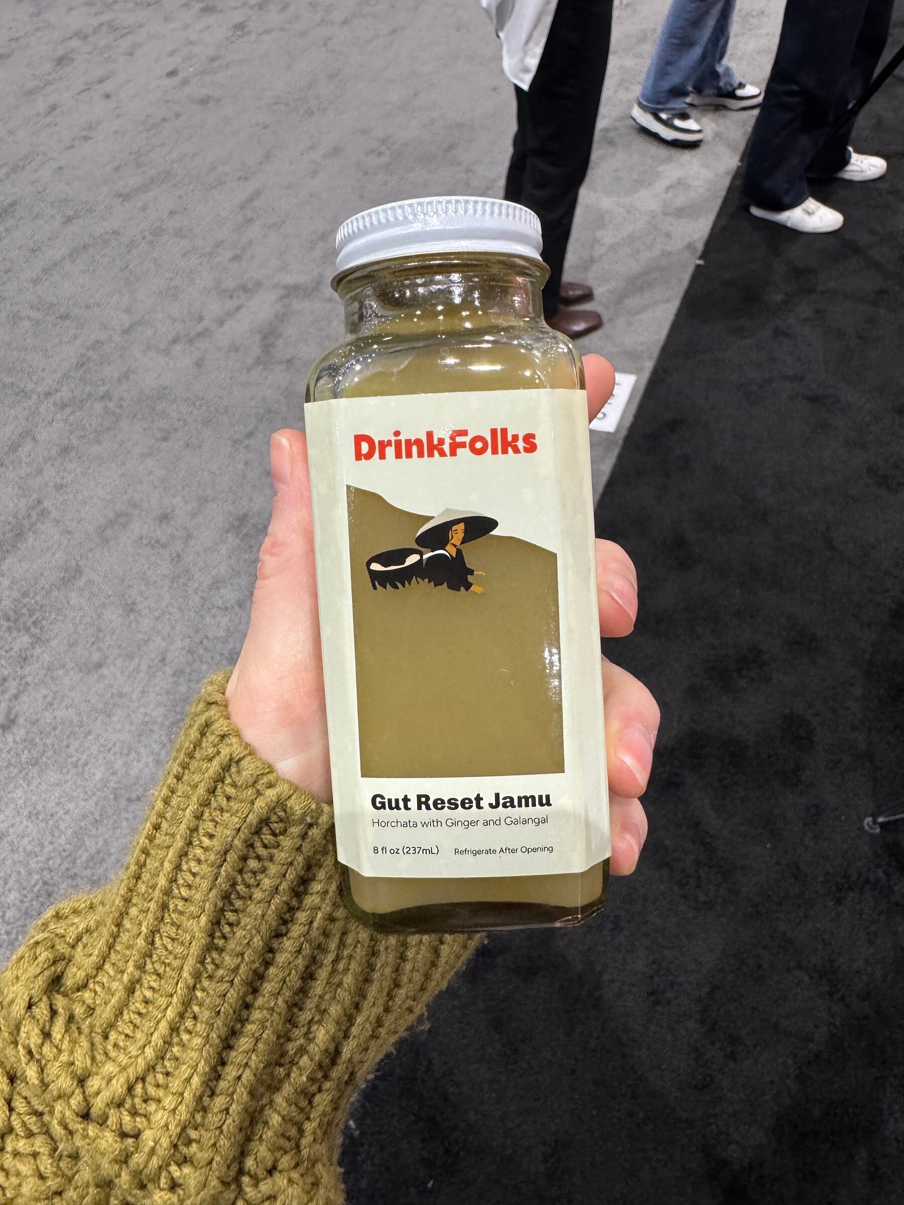 Hand holds a bottle of Drink Folks Gut Reset Jamu over the trade show carpet.