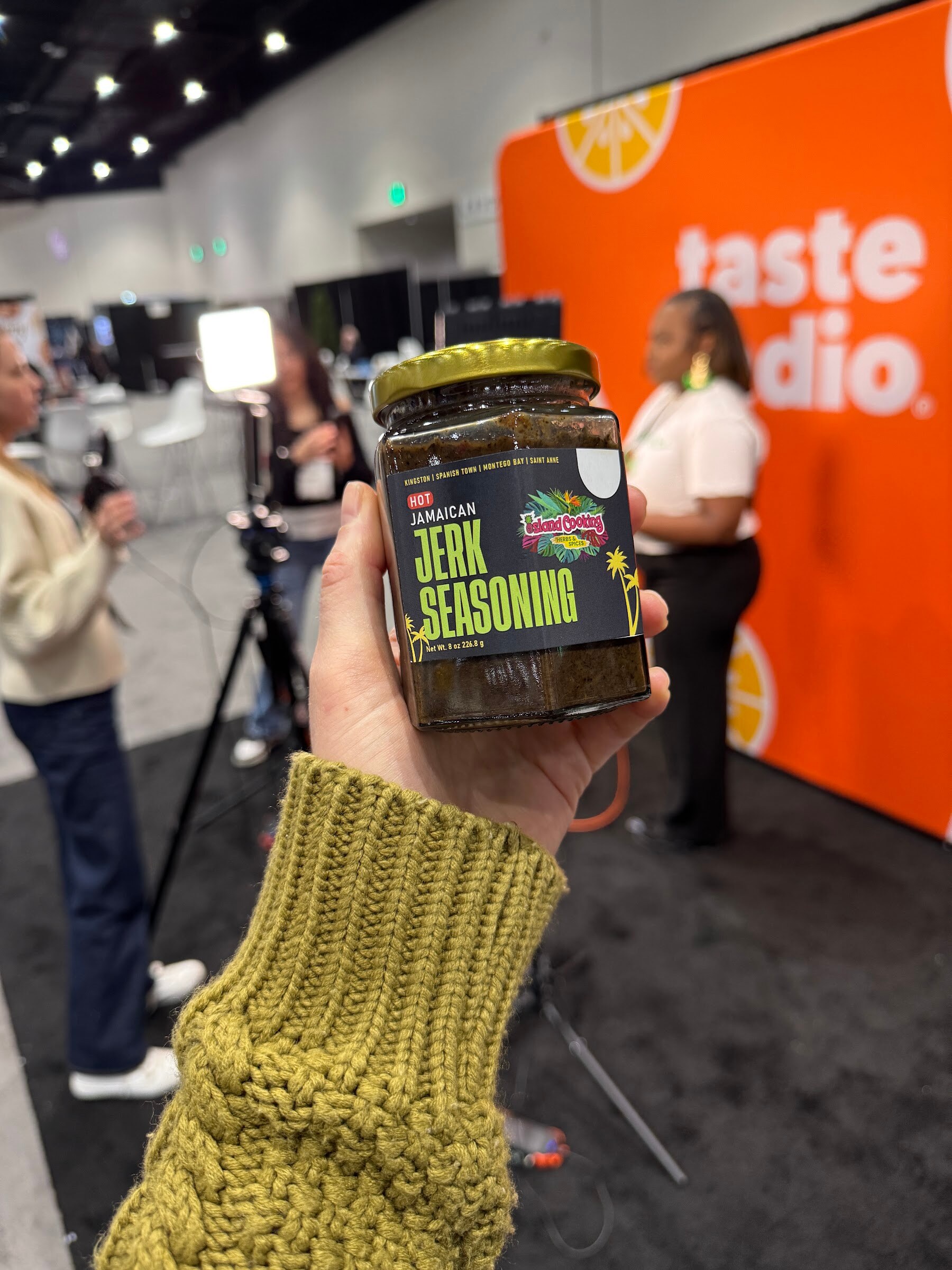 Hand holds a jar of Jamaican jerk seasoning in front of the orange Taste Radio booth.