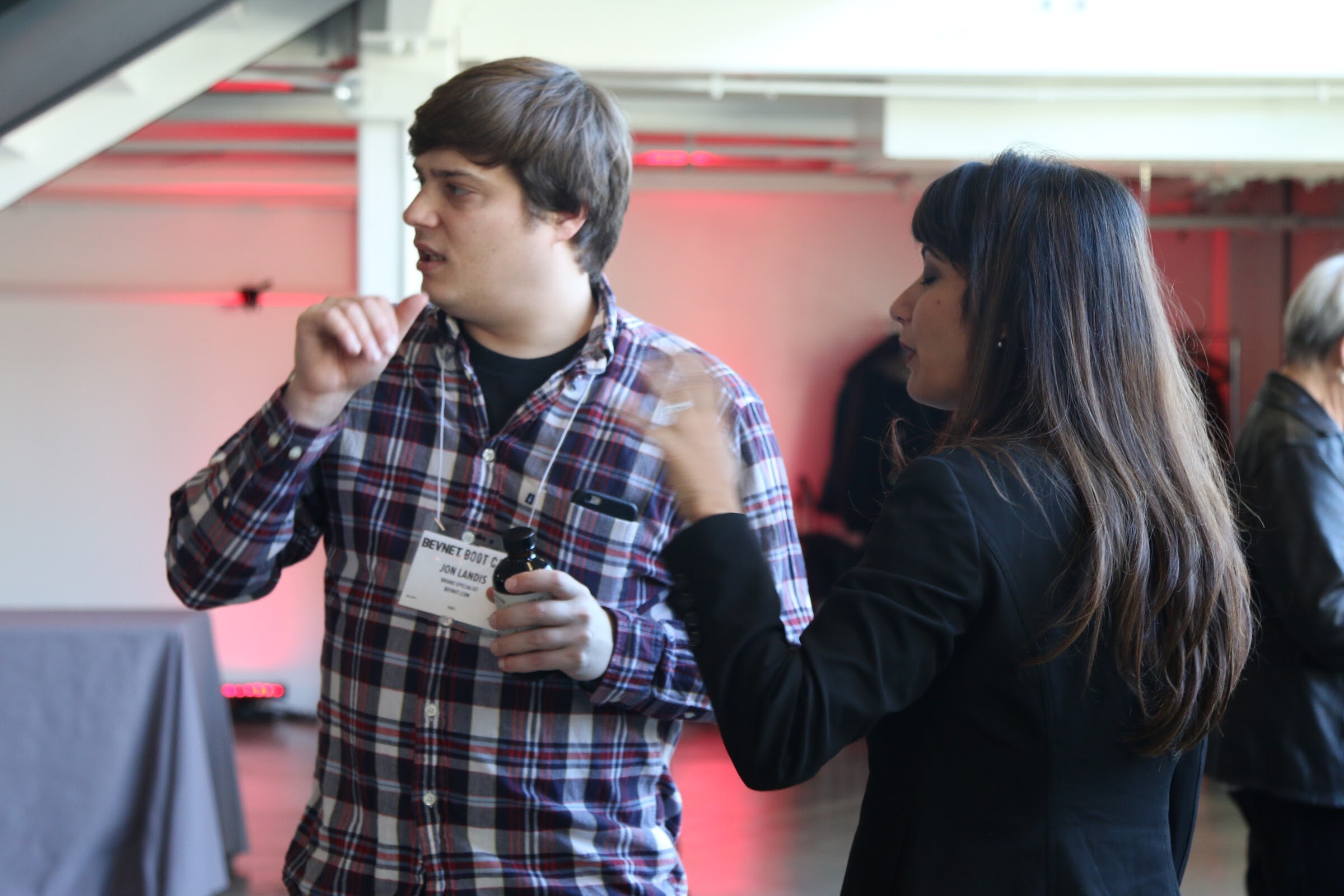 Two attendees conversing, man in plaid shirt with name badge gesturing, woman in black blazer facing him.