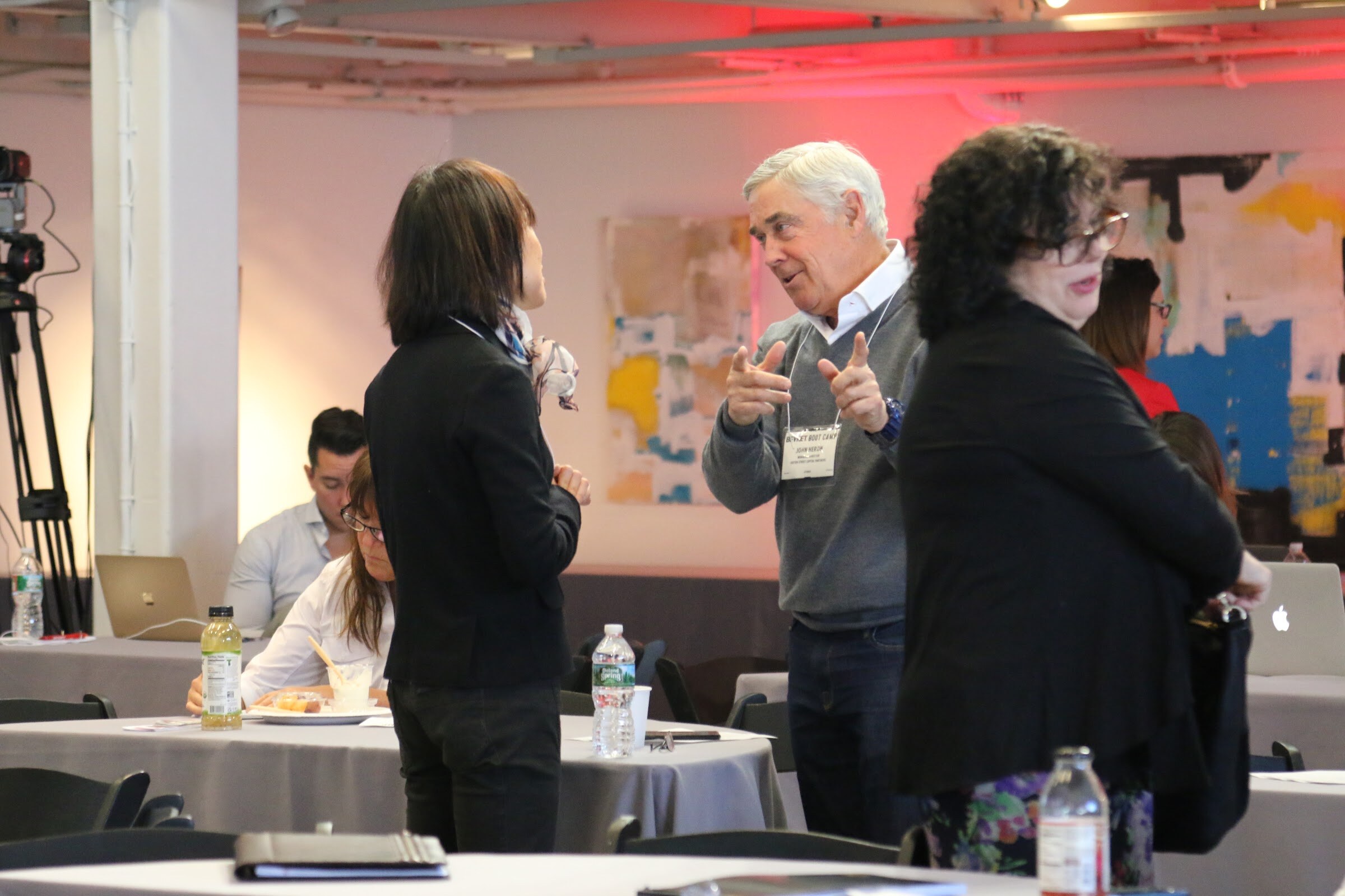 Three attendees in conversation at tables, man in grey sweater gesturing; camera tripod at left.