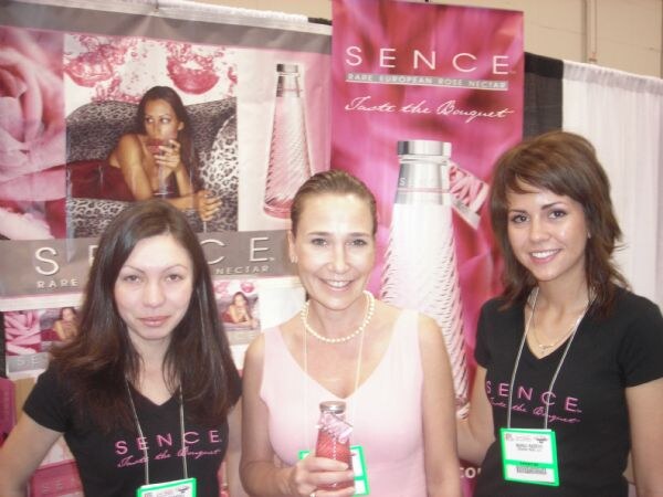 Three women stand in front of pink Sence rare European rose nectar banners; two wear black Sence shirts and the middle woman in a pink top holds a small bottle.