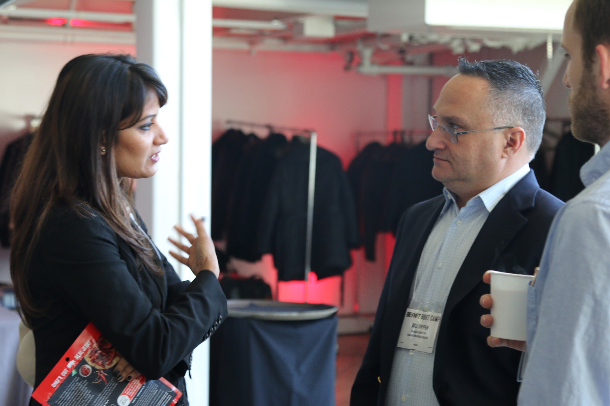 Woman talking with a man wearing a name badge and holding a coffee cup, with a pink-lit coat rack behind them.