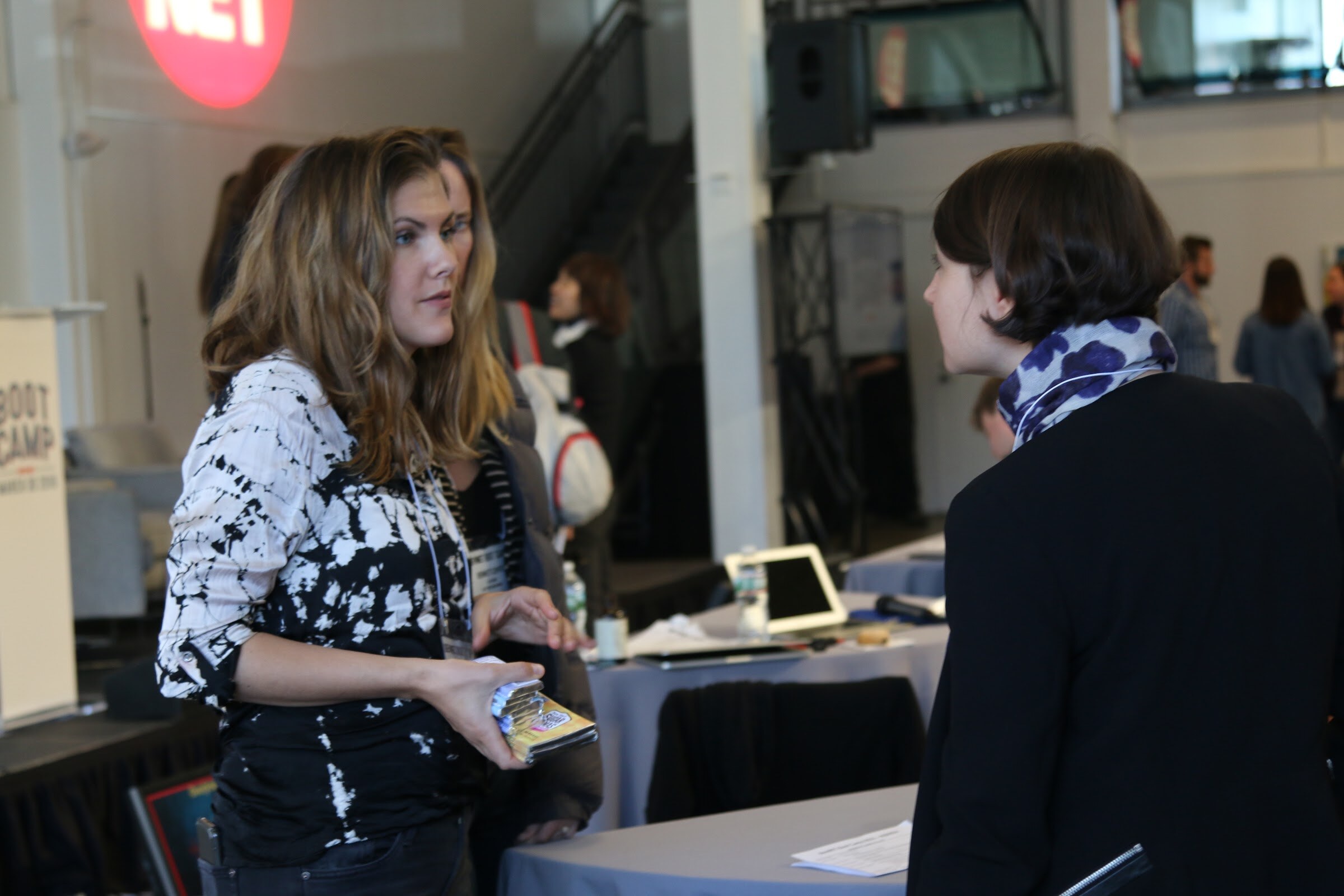 Two women talking at a high-top, Boot Camp banner left, BevNET logo wall.
