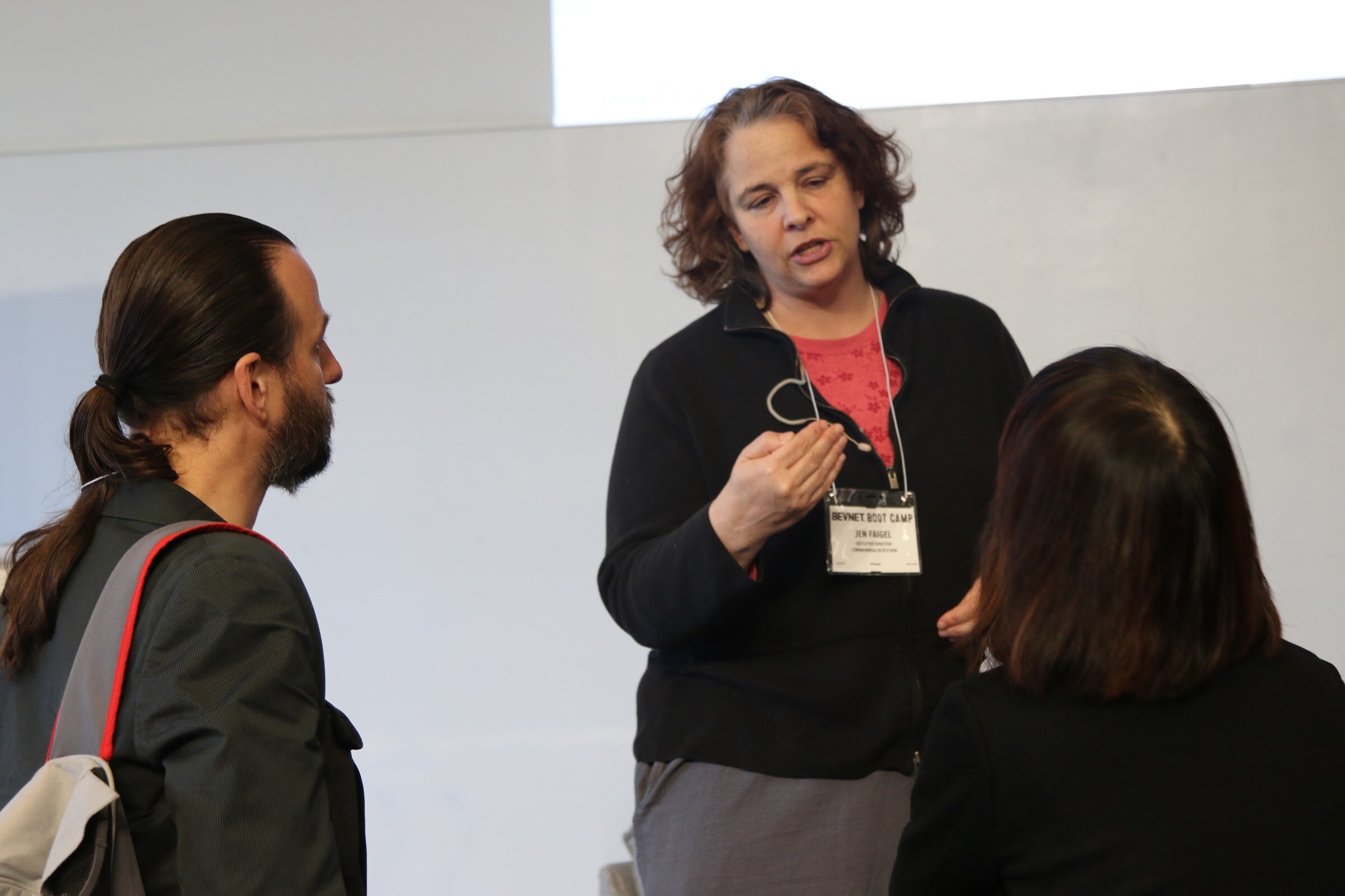 Woman speaking with two attendees, one with ponytail and red-trimmed backpack strap, name badge.