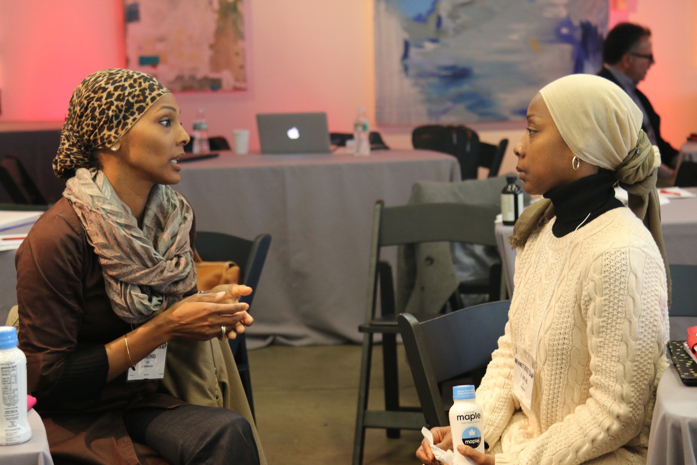 Two women in head wraps talking, Maple bottles in foreground, pink-lit art behind.
