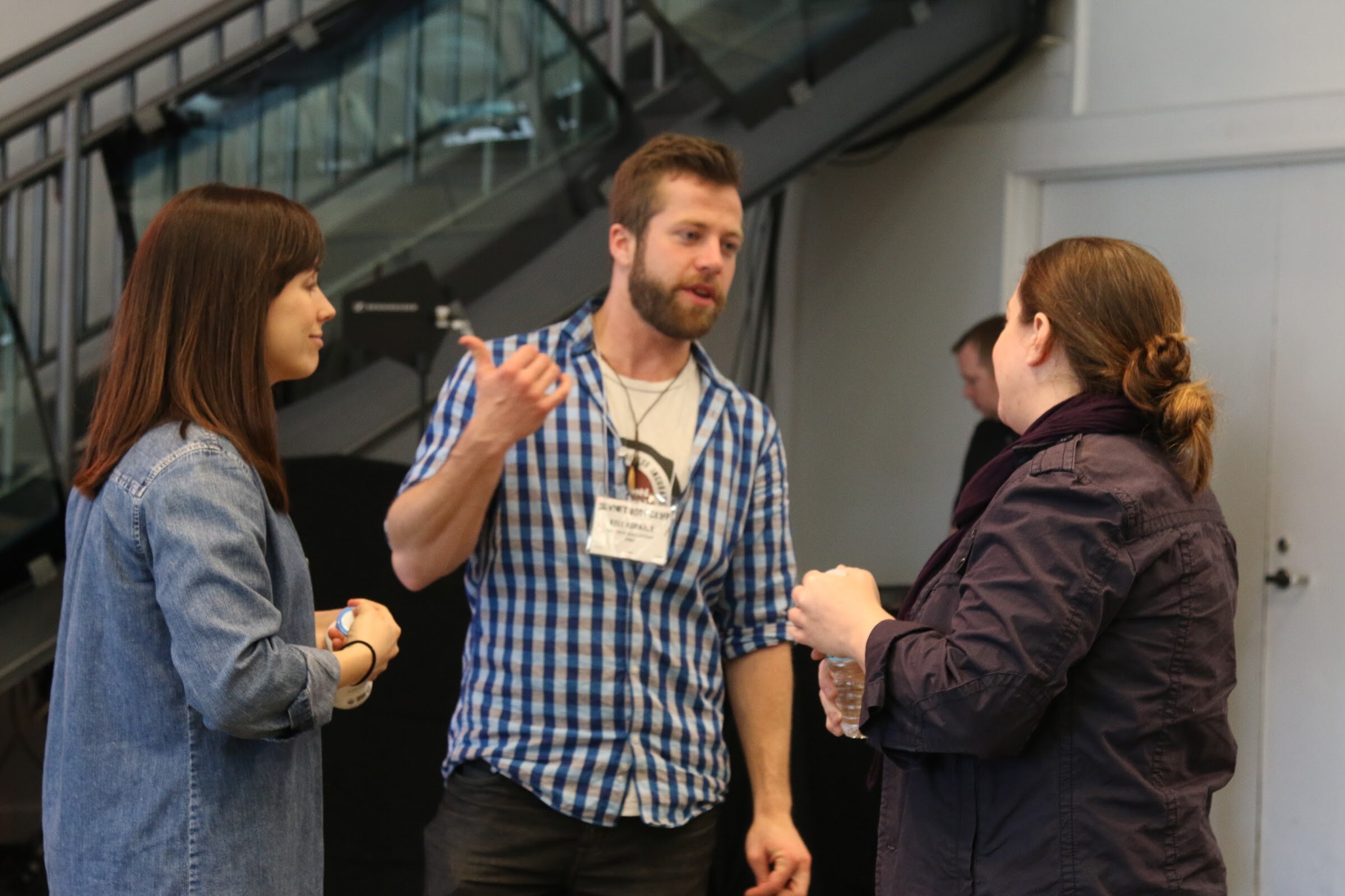 Man in blue plaid with name badge gesturing to two women, stairs behind.