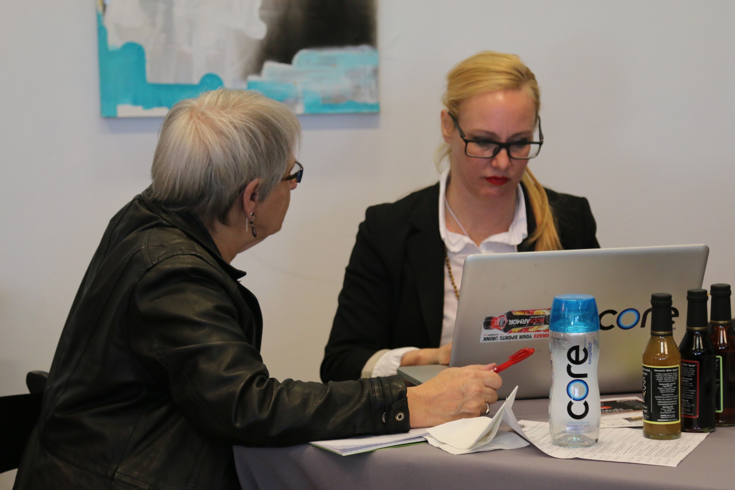 Two women at table, one at Core laptop with Core Hydration bottle and bottles of vinegar/syrup, other leaning in.