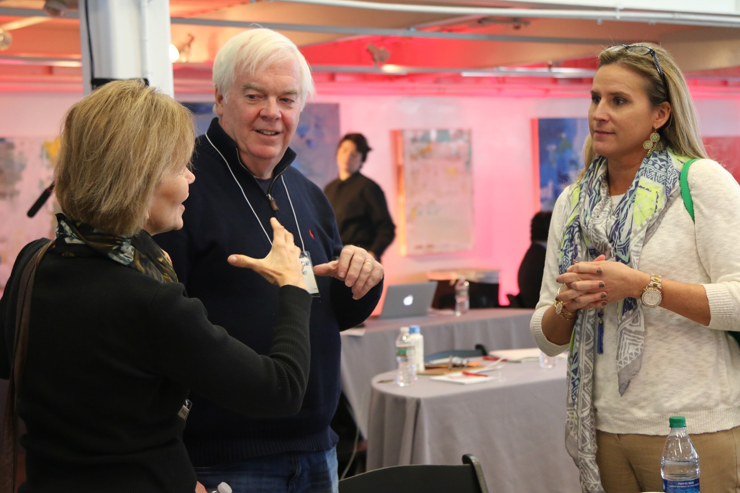 Three attendees talk, pink-lit room, older man gesturing.