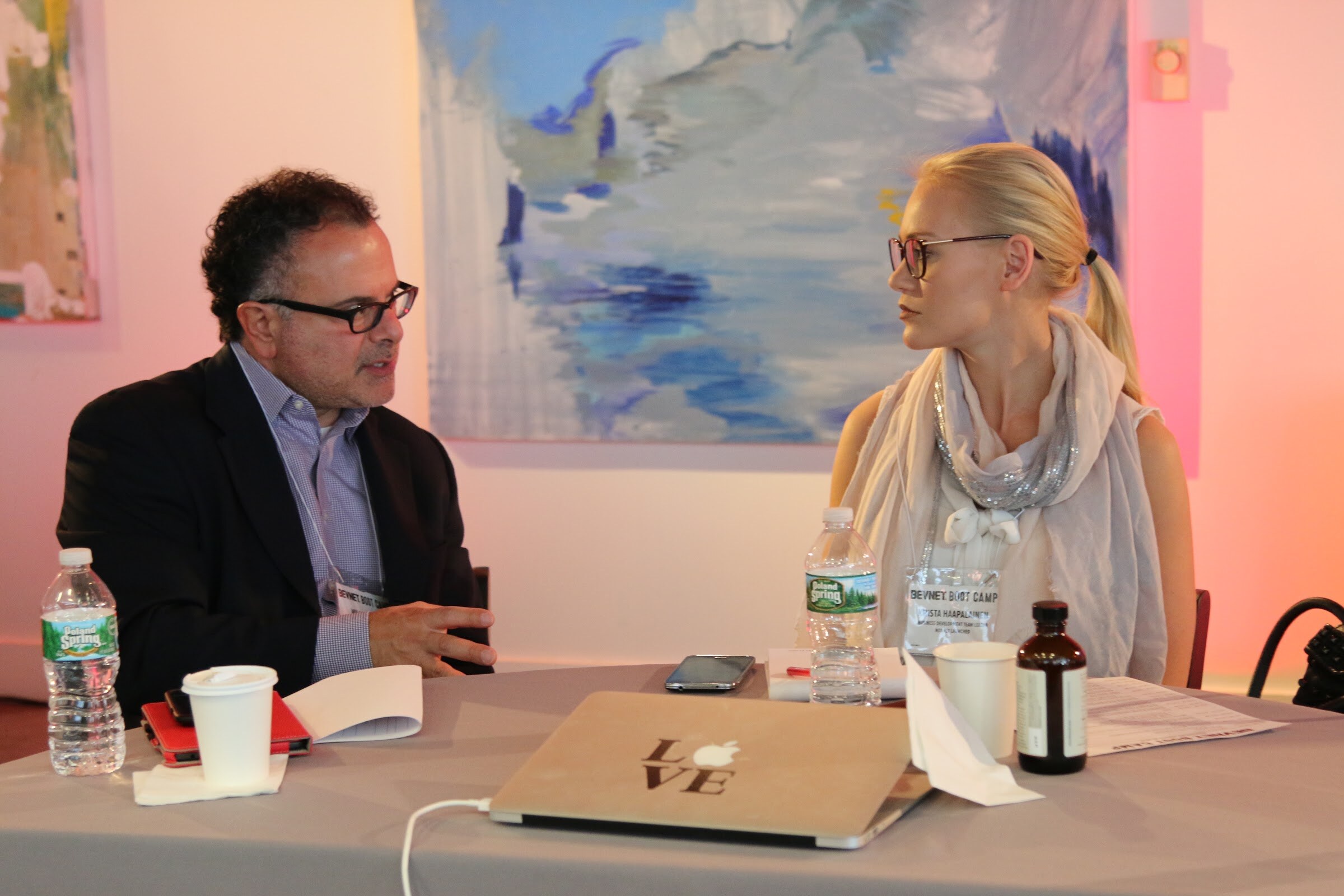 Man and woman talk at table, laptop with LOVE sticker, Poland Spring bottles.