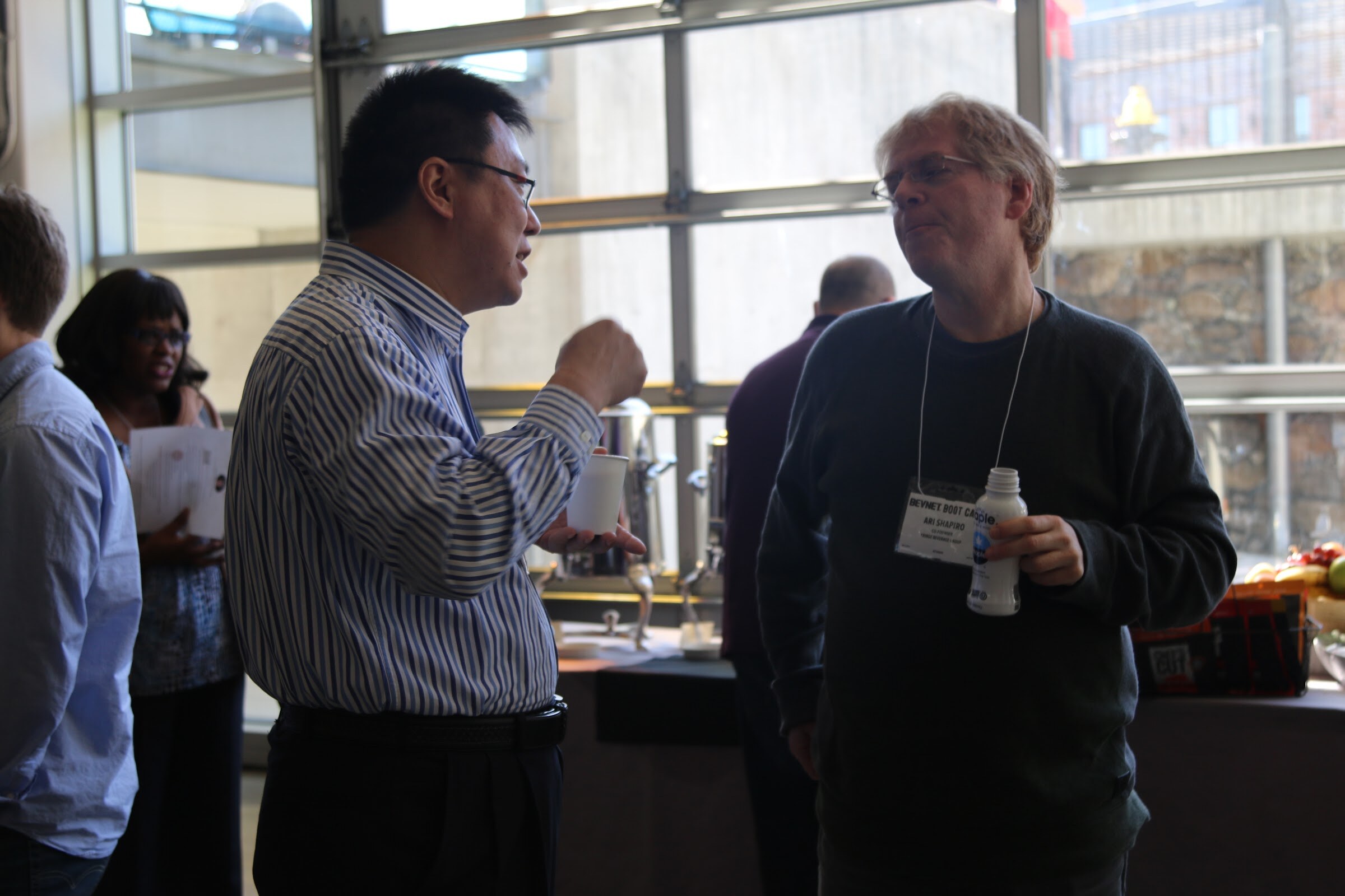 Two men chatting near coffee urns in front of glass doors, one holding Maple bottle.