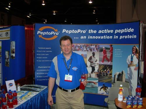 A man in a blue polo shirt with a lanyard badge holds a red bottle in front of a blue PeptoPro banner reading "the active peptide... an innovation in protein"; bottles sit on a table nearby.