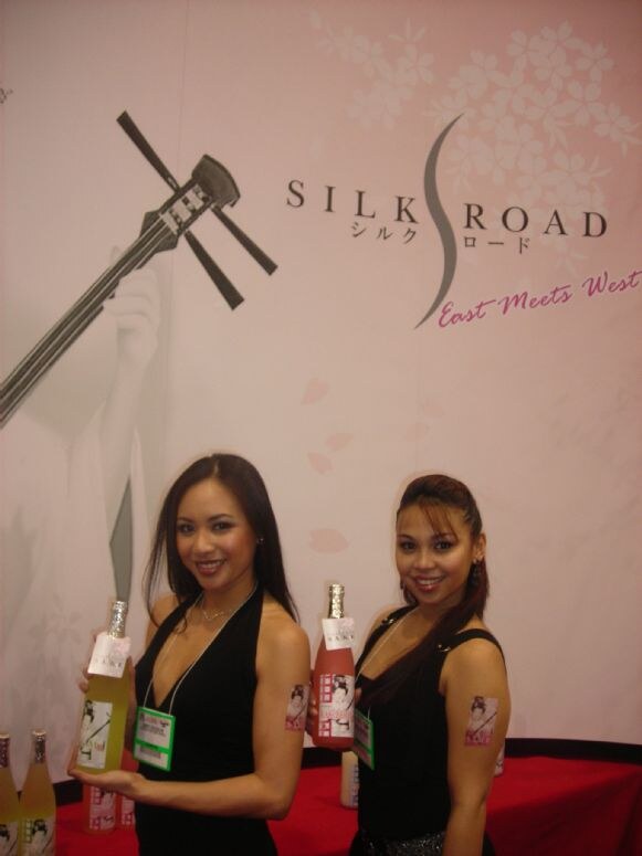 Two women in black dresses hold bottles in front of a pink Silk Road backdrop reading East Meets West, with more bottles on a red-draped table.