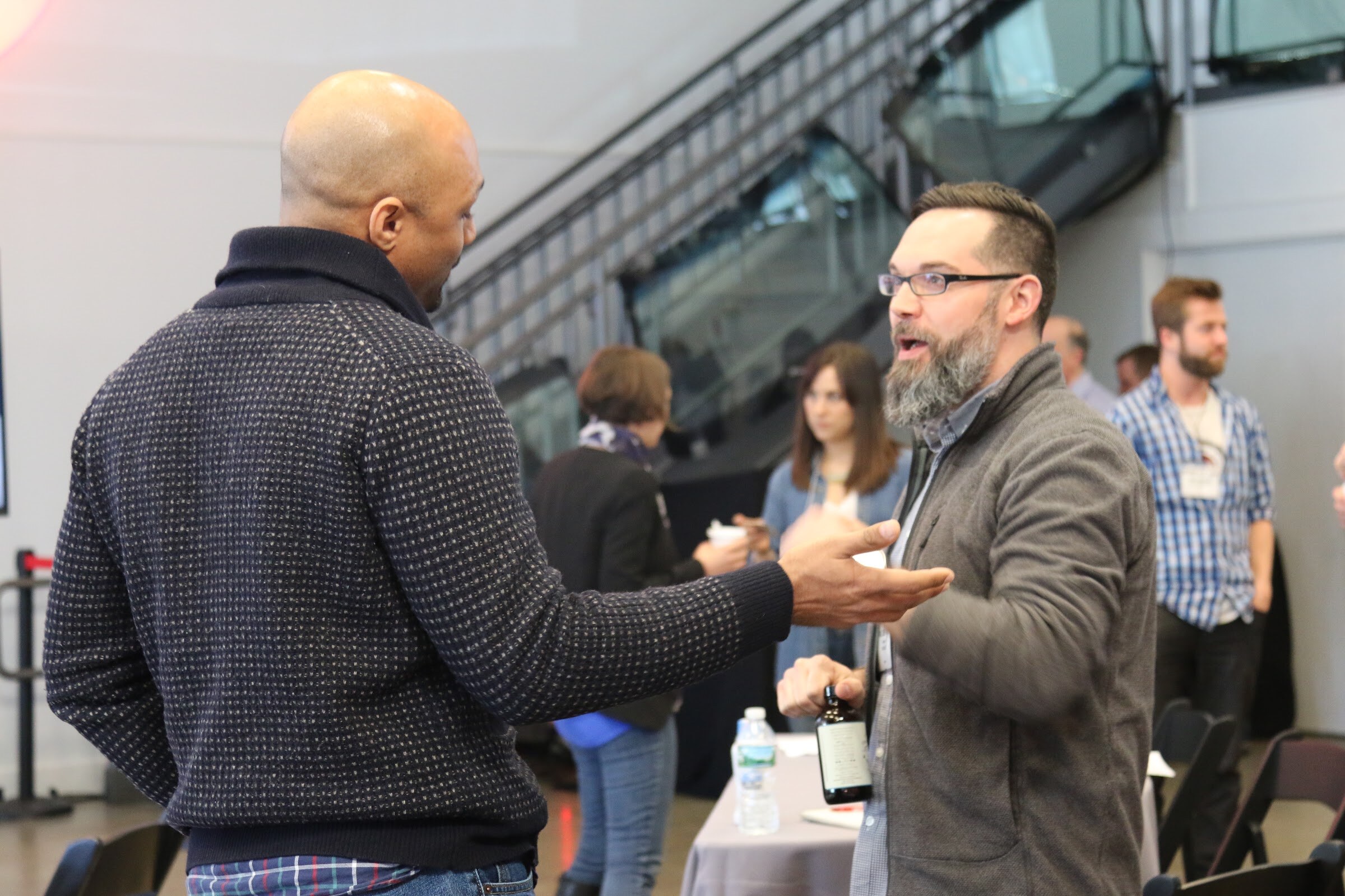 Two men conversing, one bald in speckled sweater with back to camera, bearded man answering.