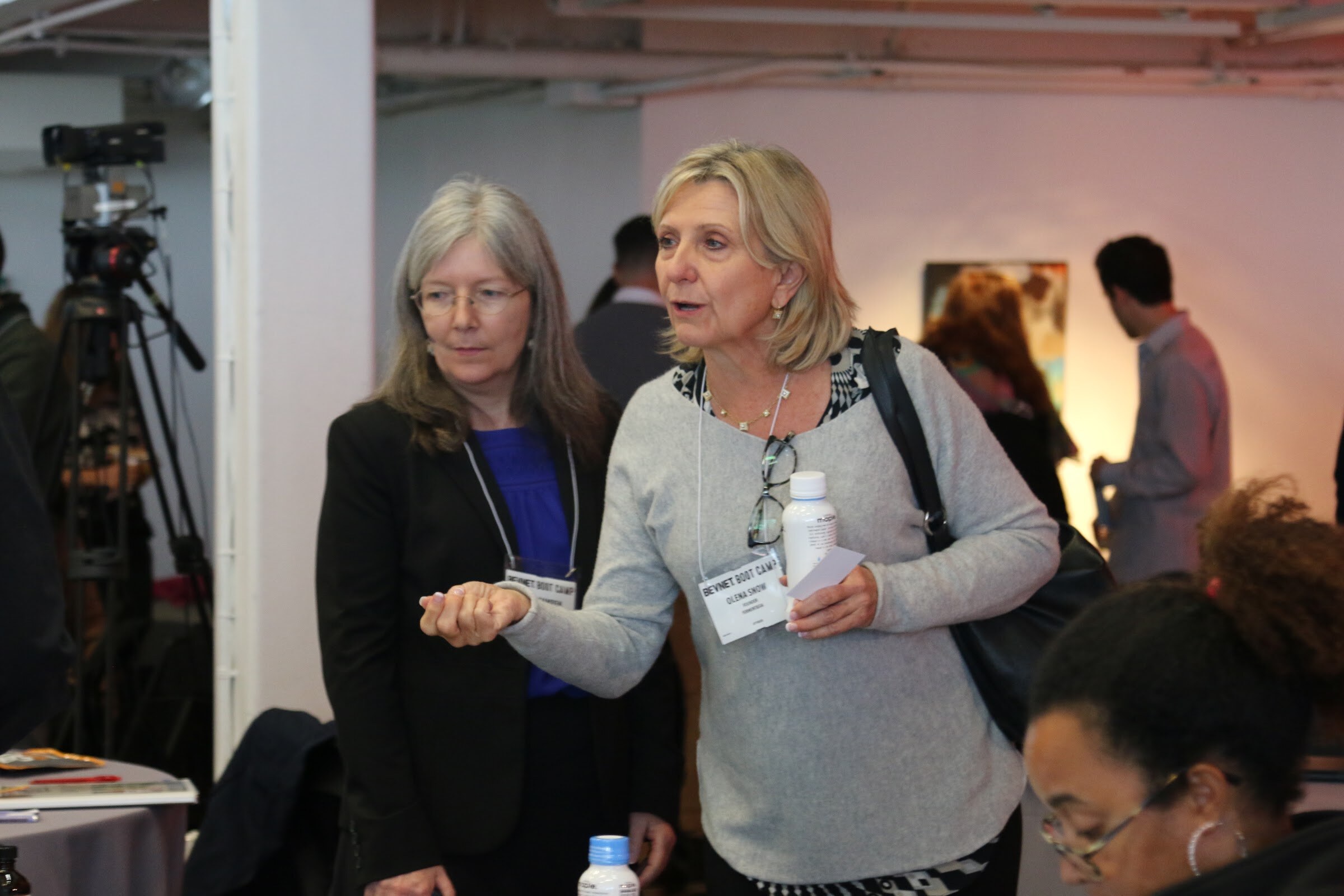 Two women in conversation at a table, camera on tripod at left, one holding Maple bottle.