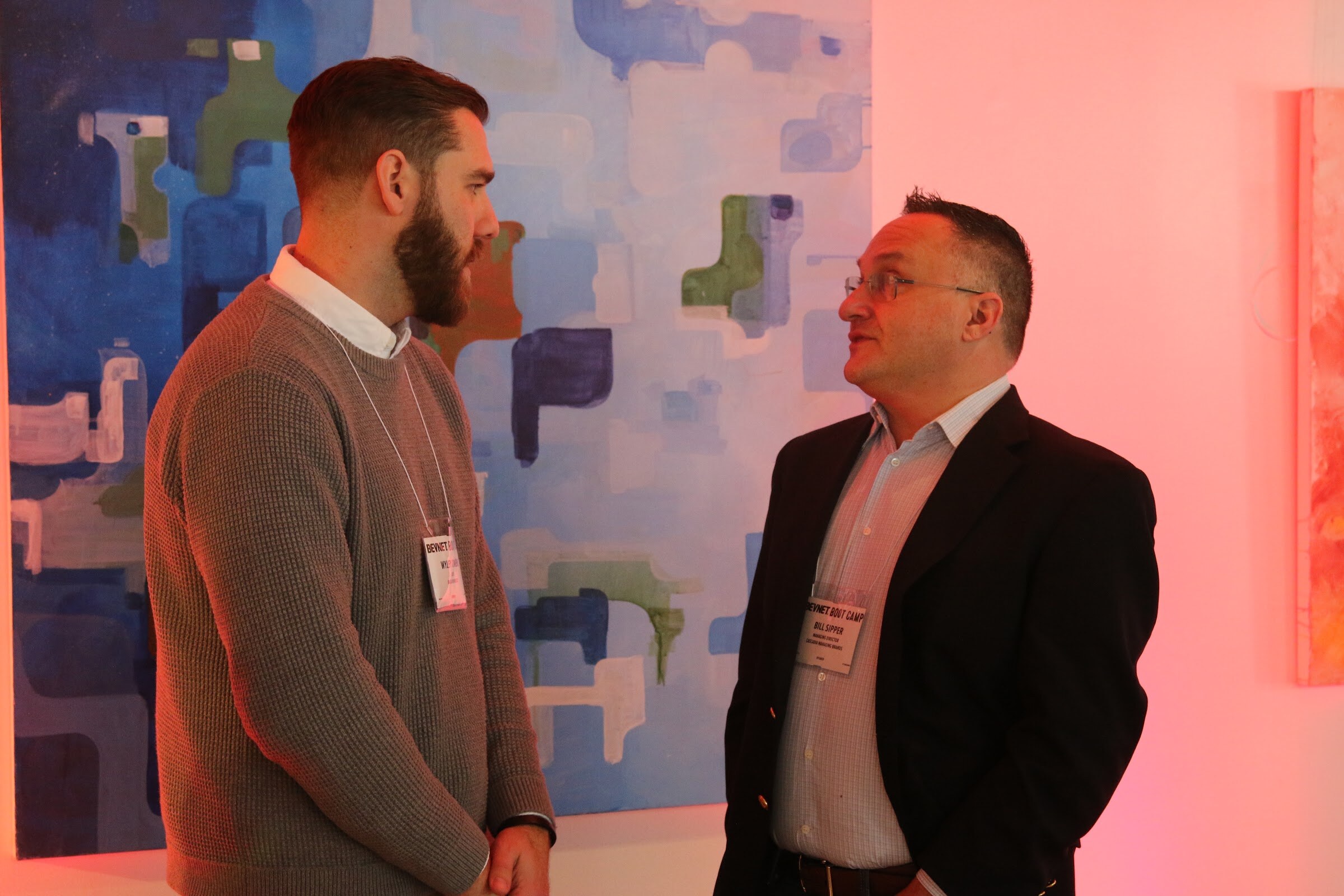Two men talking in front of abstract painting in pink light, name badges.