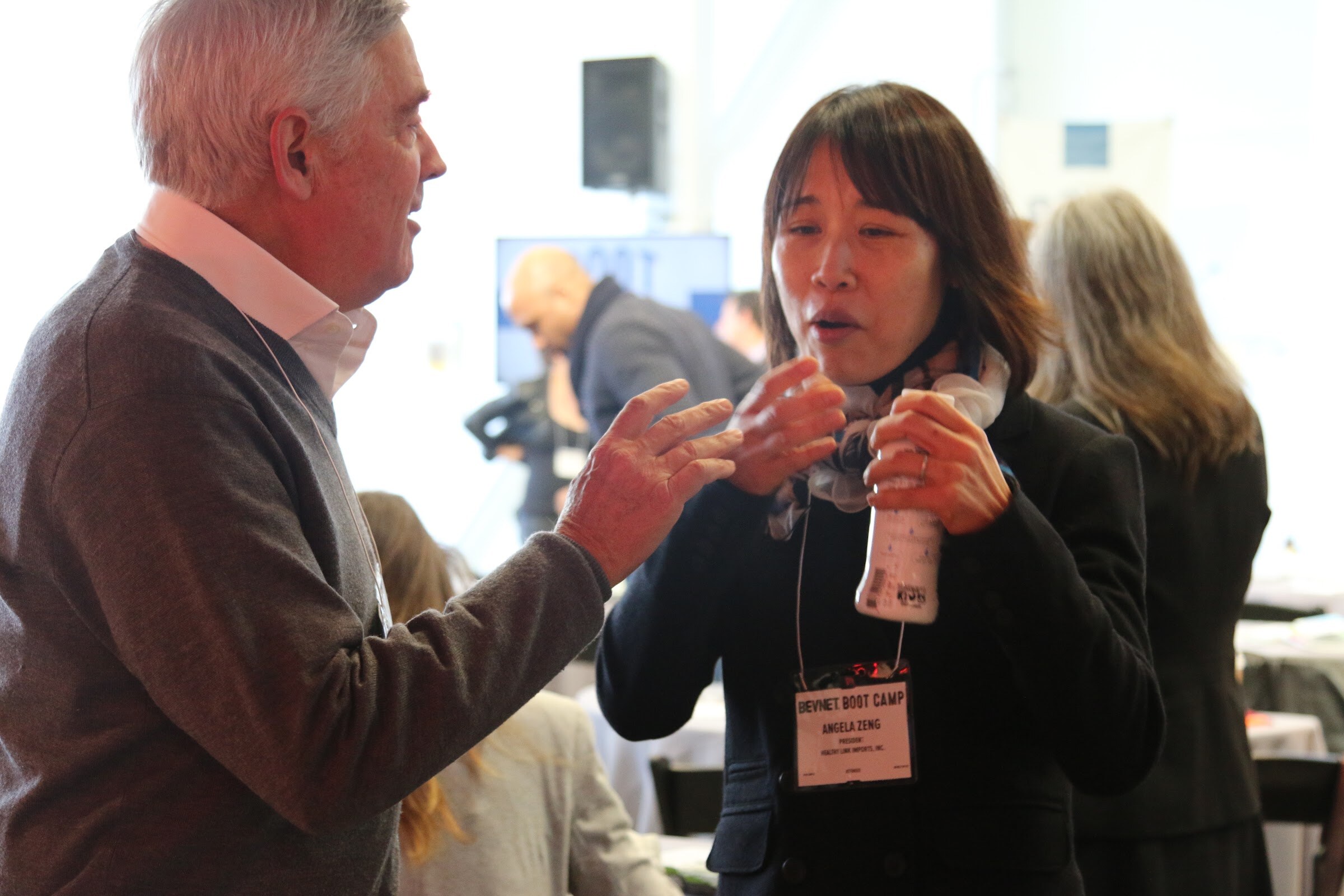 Man in grey sweater gestures while woman in dark jacket holding bottle speaks, name badges.