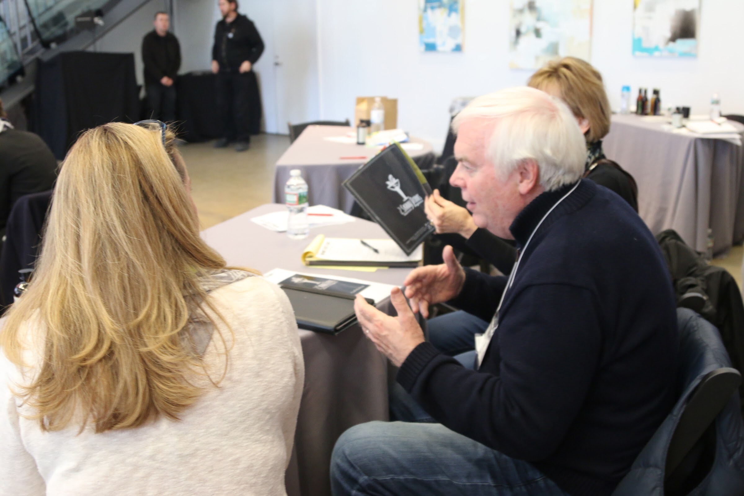 Man with white hair talks to woman at table, others seated nearby, Boot Camp attendees at tables.