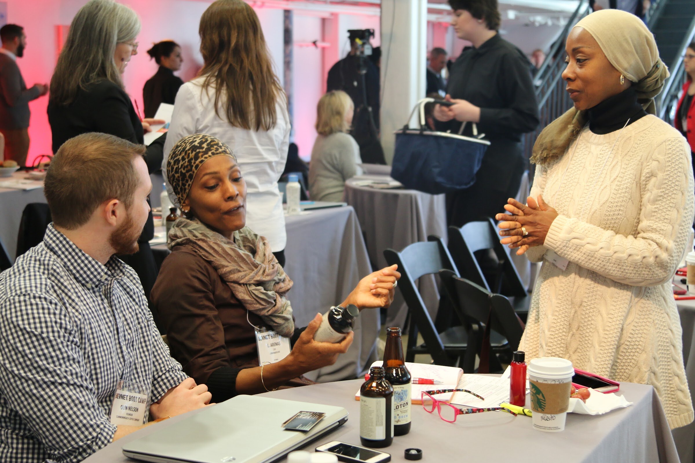 Attendees chatting at a table: woman holds a bottle, other with Starbucks cup, bottles of brew on table.