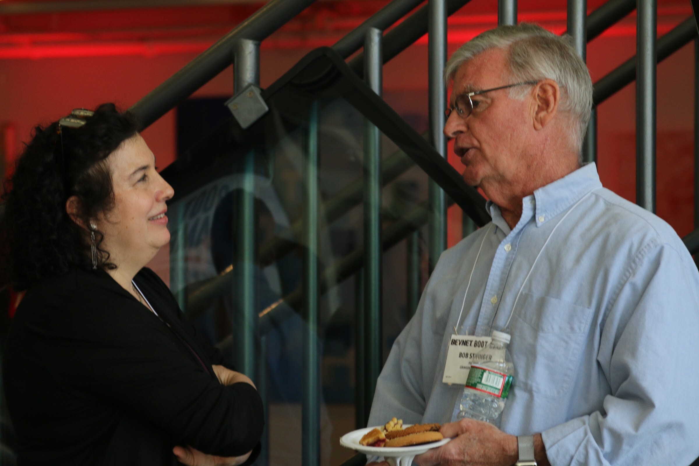Two attendees chatting by a glass staircase, one holding a plate of cookies and a water bottle.