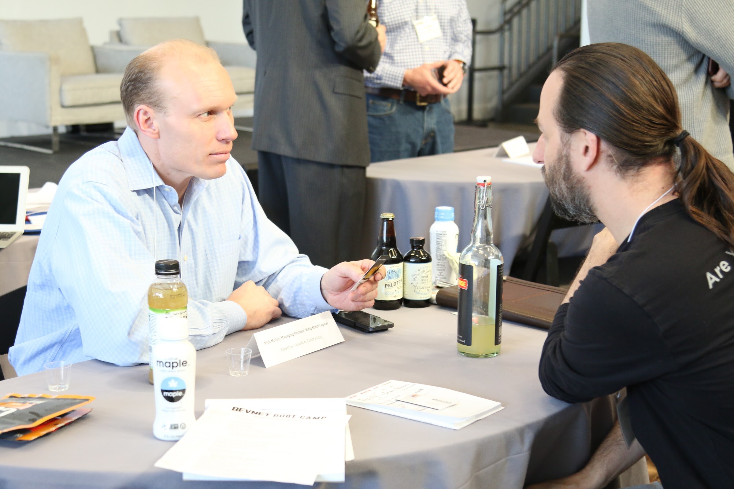 Man in blue shirt holds a card across table from bearded man with ponytail, Maple bottle and craft beer bottles on table.