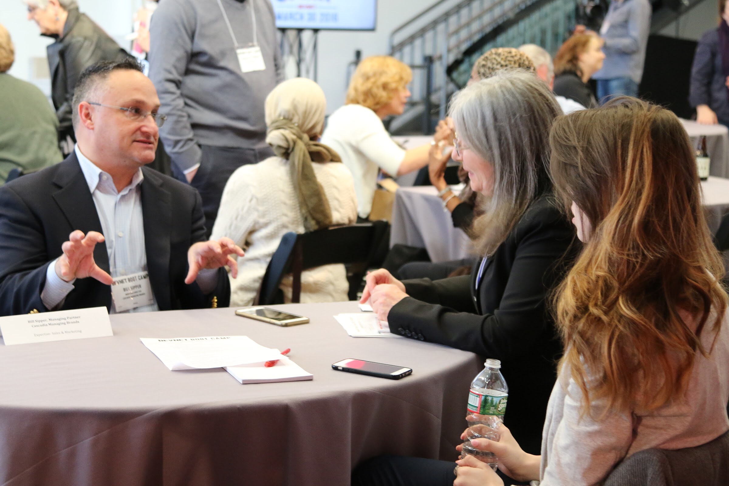 Man in blazer gesturing at a table meeting with two women, name placard, phones and papers on table.
