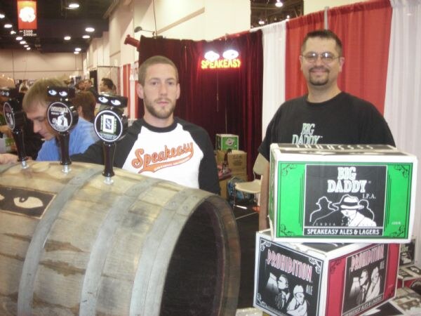 Two men stand by a wooden barrel with taps and cartons of Big Daddy IPA and Prohibition Ale at a Speakeasy booth; a third man sits partly visible at left.