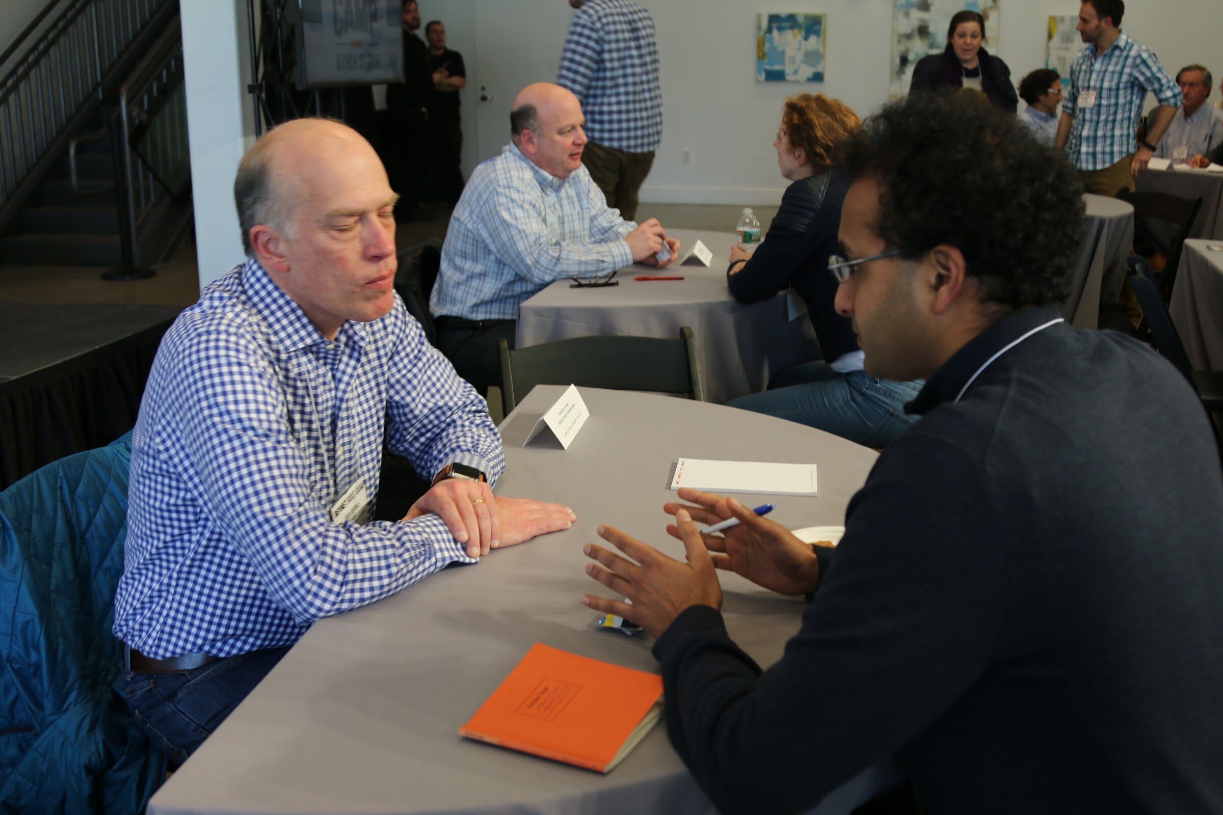 One-on-one meeting: two men talking across a round table, others meeting in background.