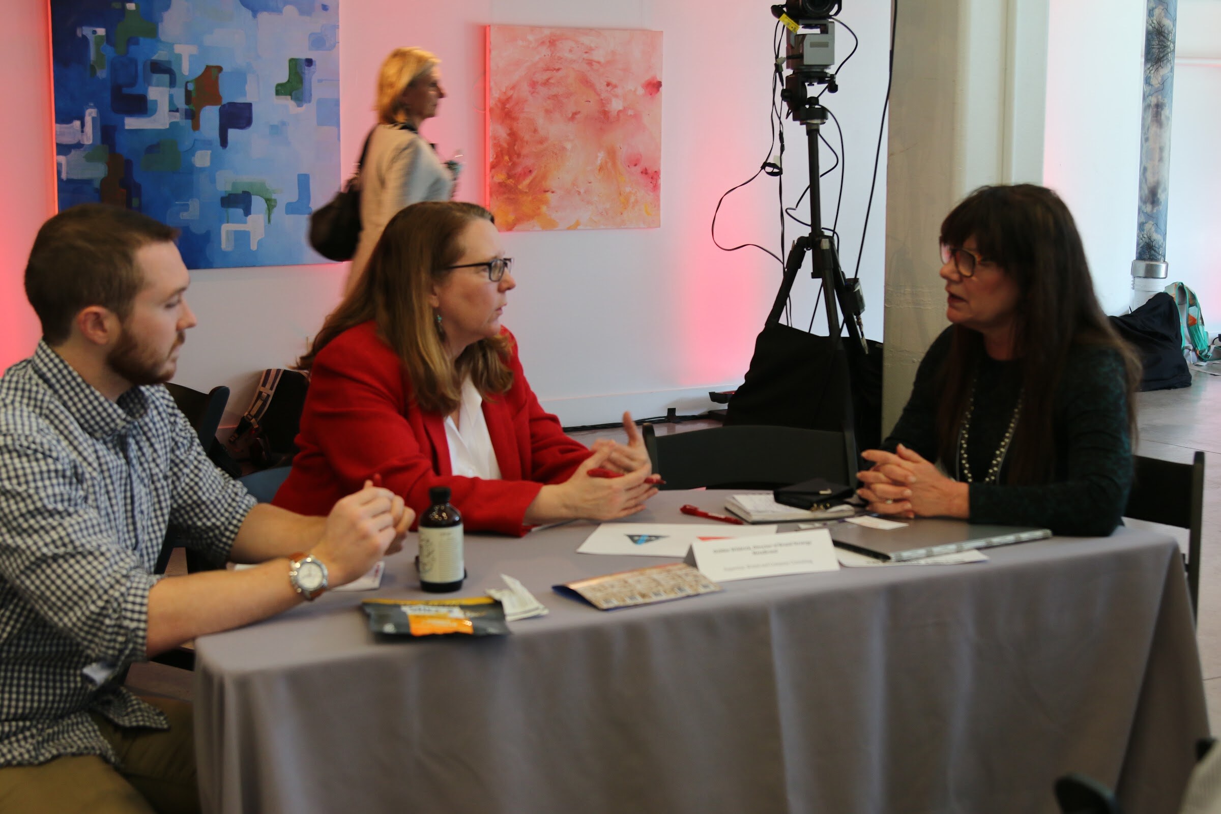 Three people in a table meeting in front of pink-lit abstract paintings, camera tripod behind.