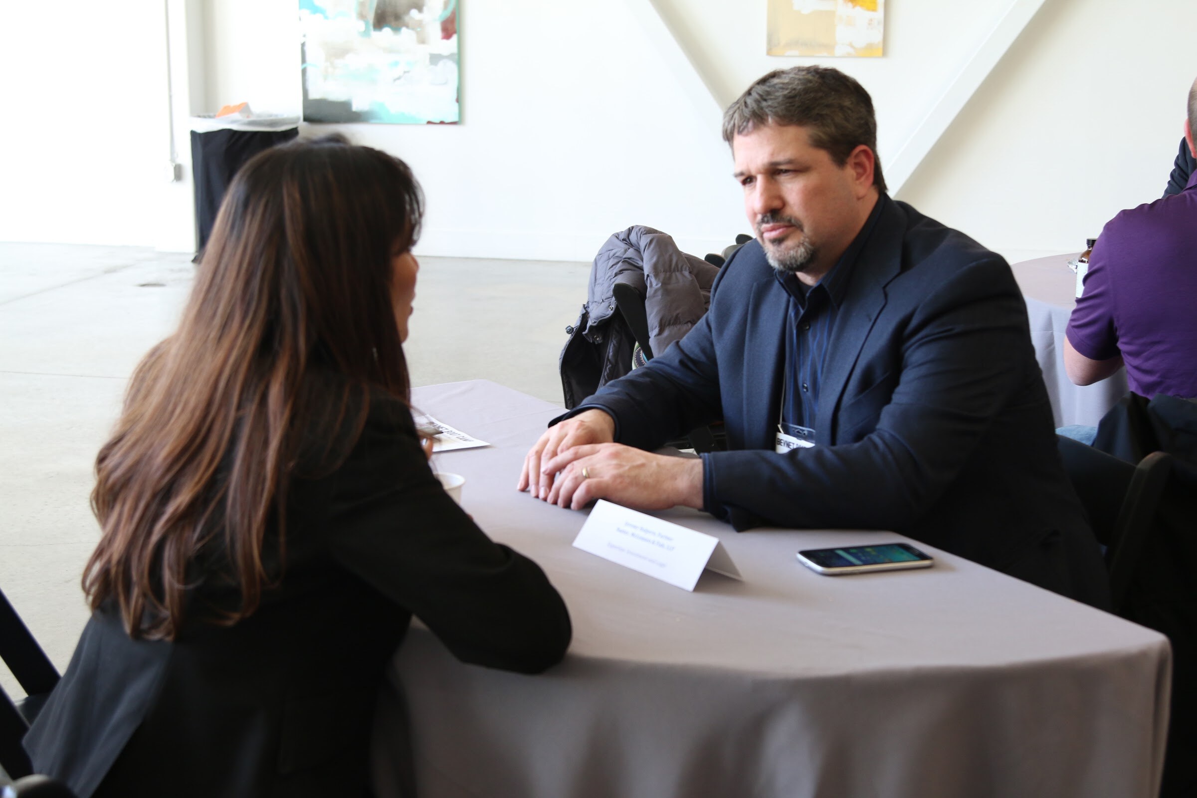 Man in navy blazer listens to woman across a table with name placard.