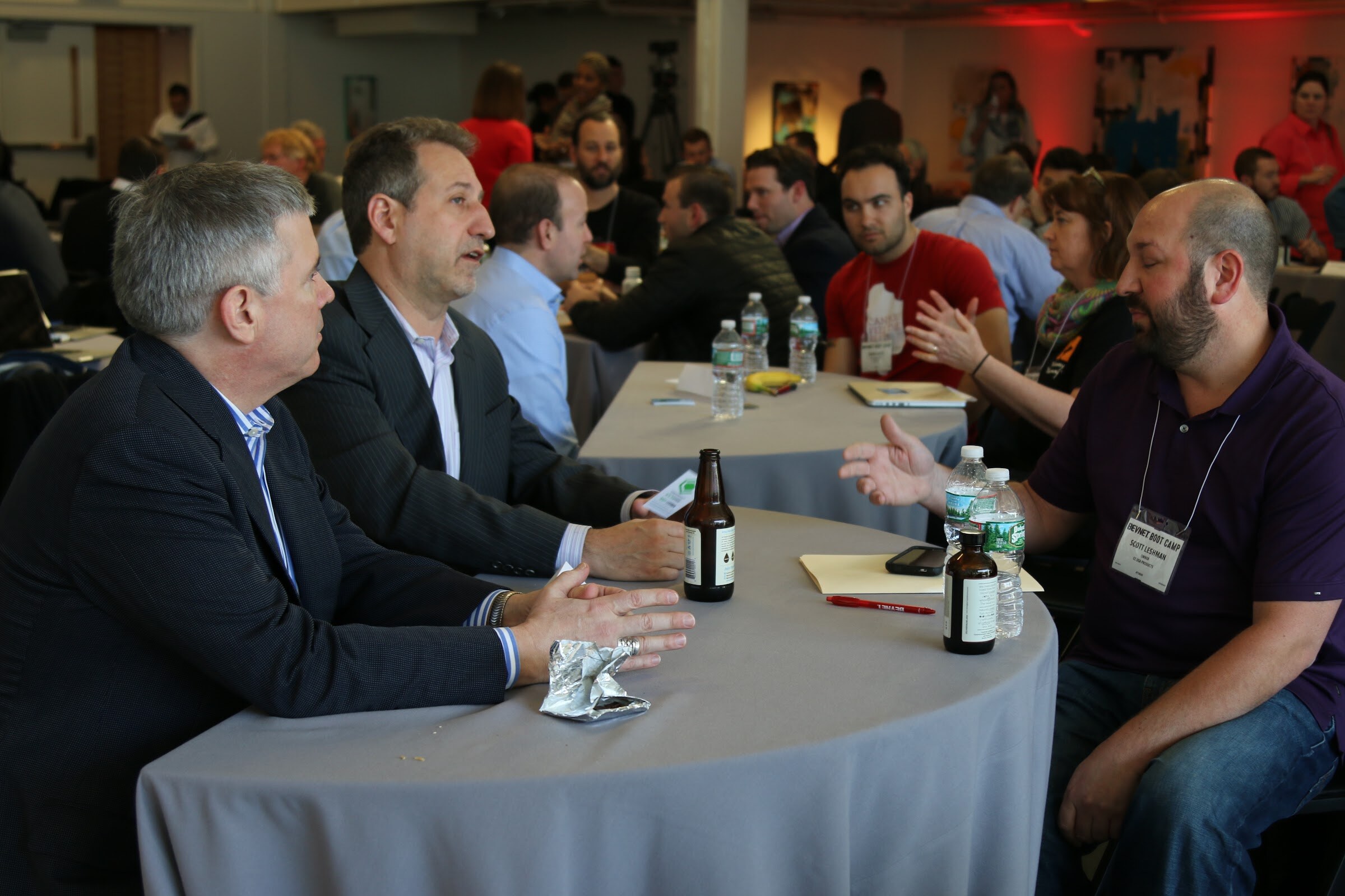 Row of meetings at a long curved table, attendees in conversation with beers, notepads.