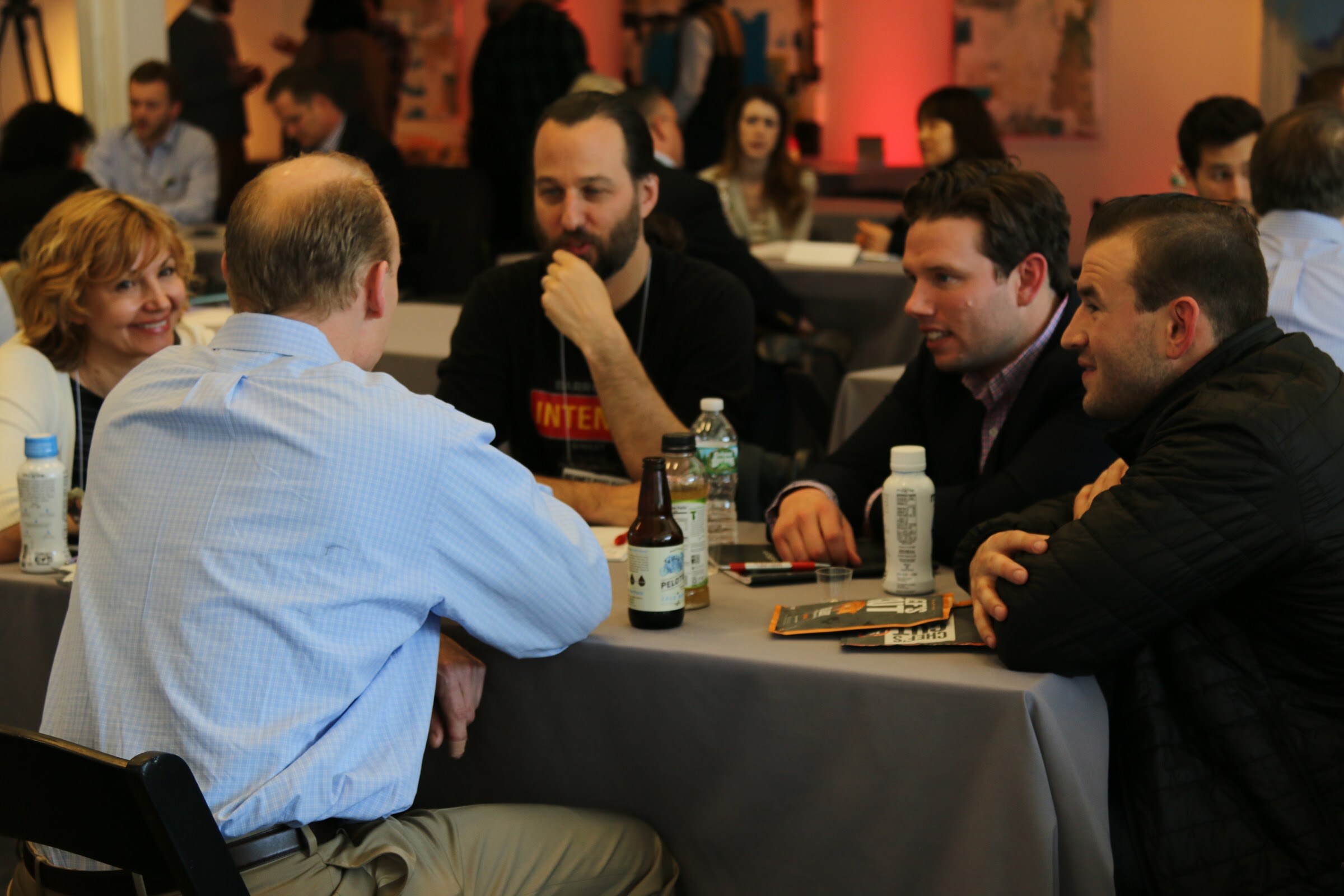 Table meeting: three men and a woman in conversation, beers and Maple bottle, name badges.
