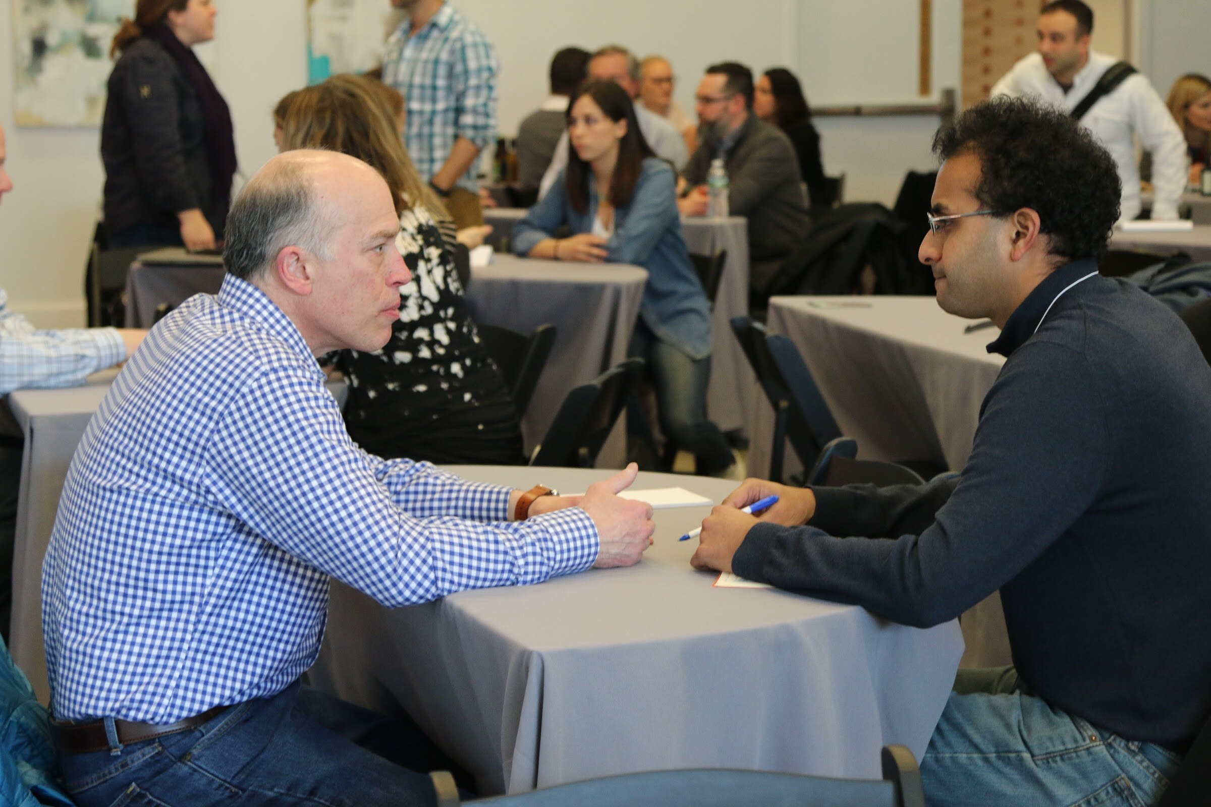 Two men in a one-on-one conversation across a round table, other attendees meeting behind.