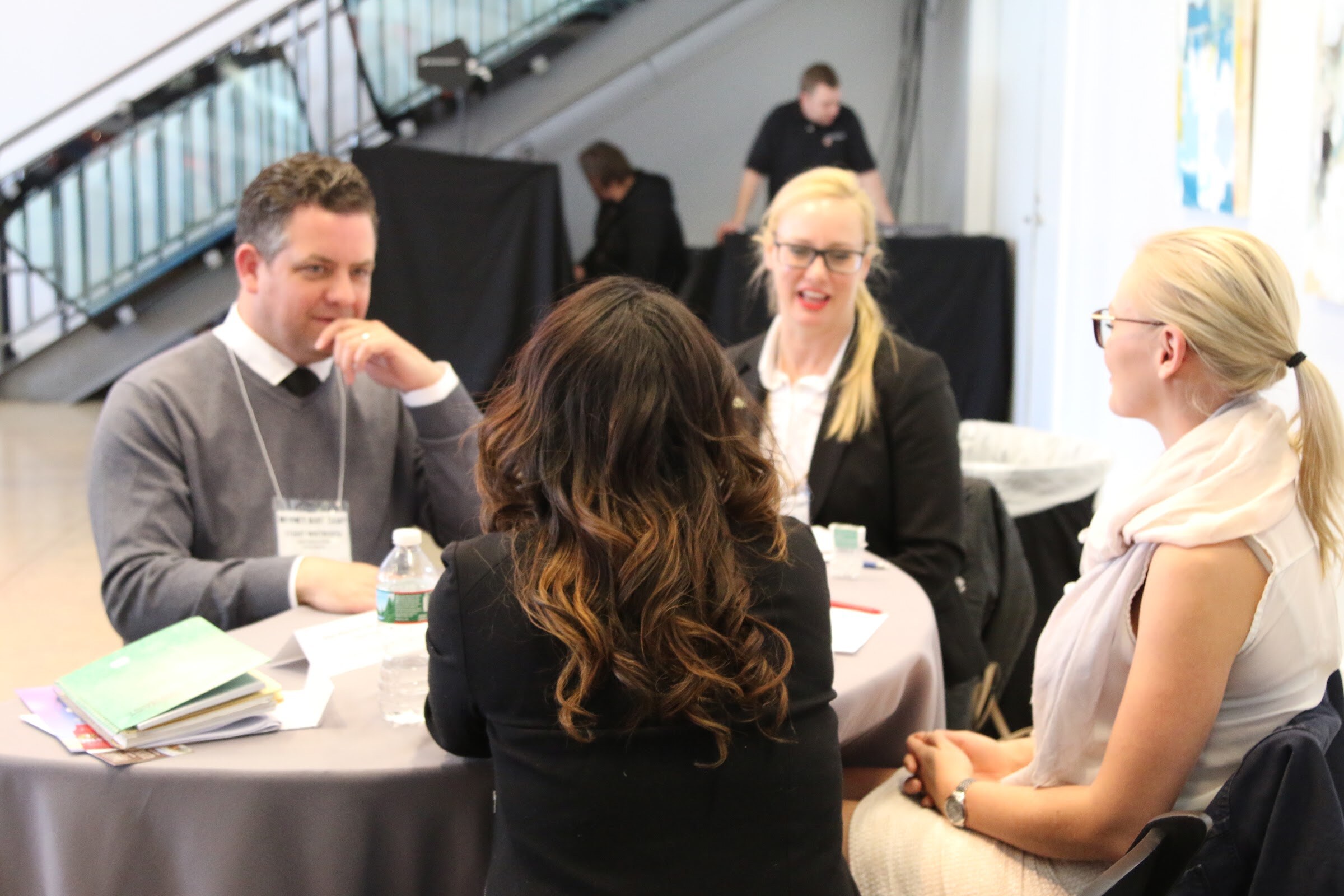 Meeting at a round table, man in grey sweater lecturing to two women facing, background staff.