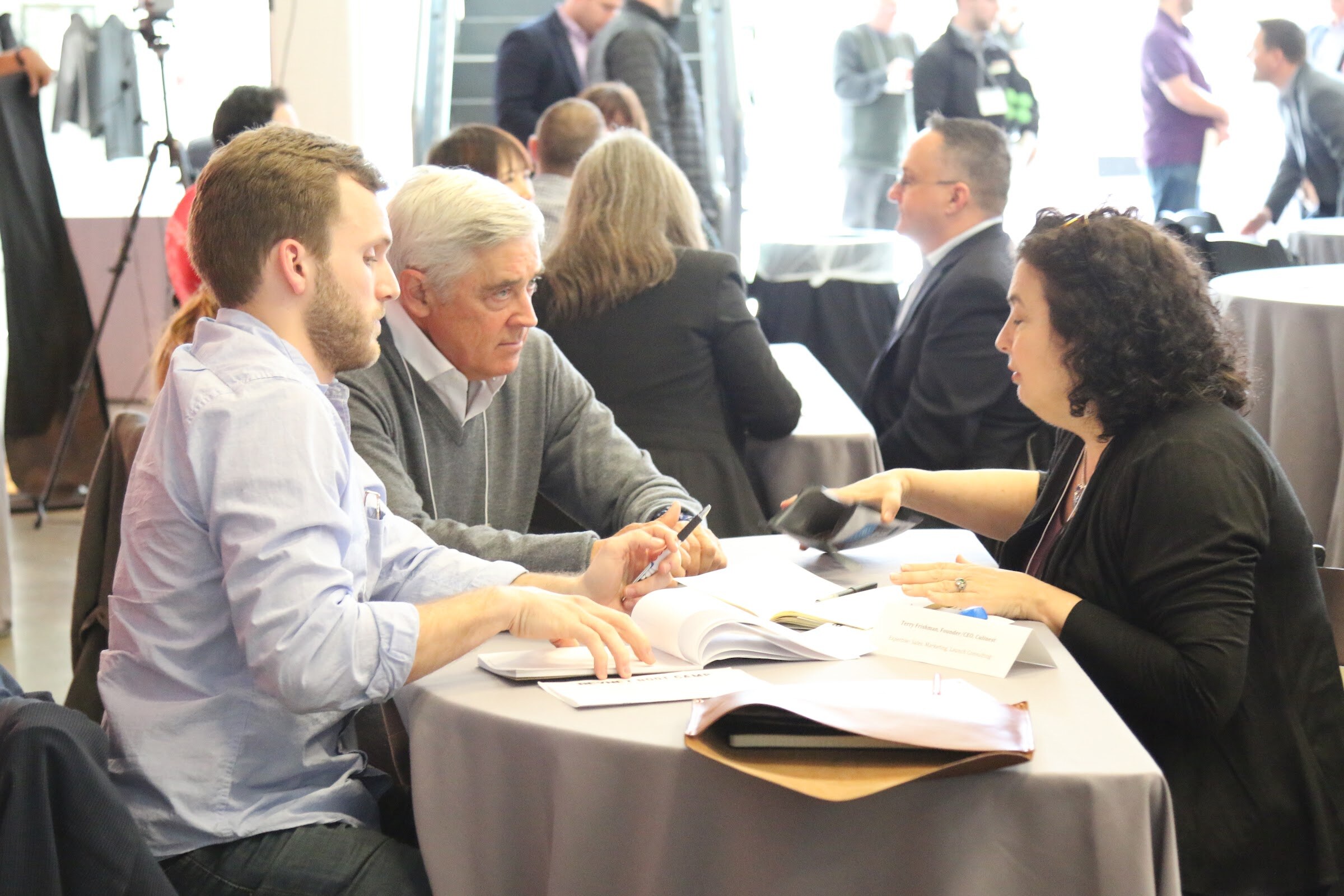 Meeting at a round table: older man and younger man review papers with woman, other meetings behind.
